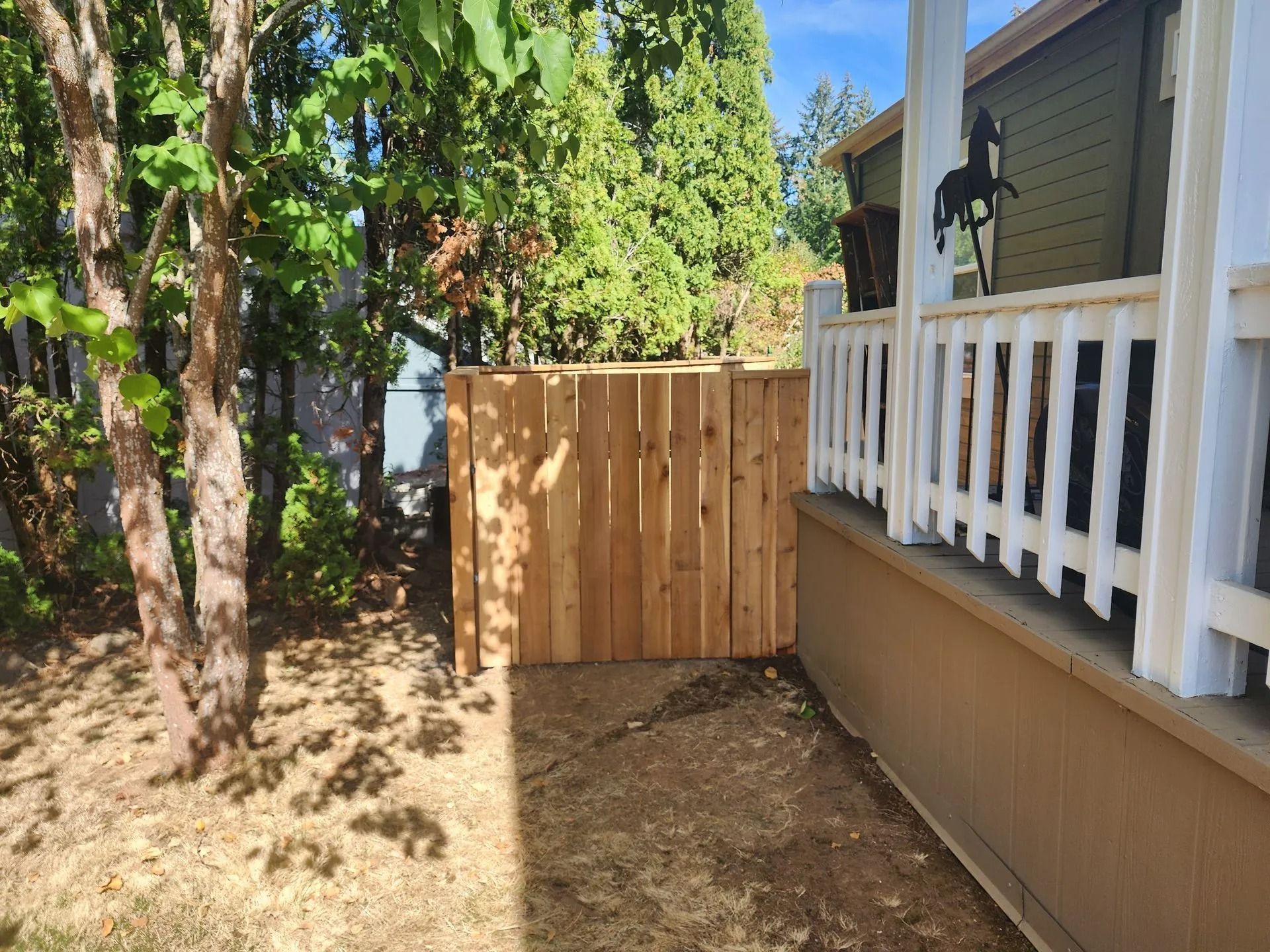 A light-colored wooden privacy fence stands between a tree-filled yard and the side of a house with a white porch railing.
