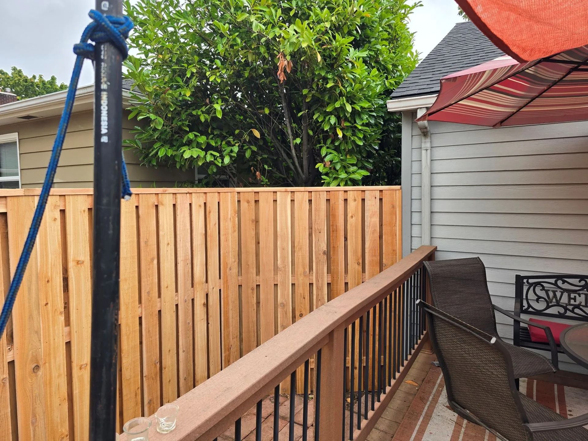 A wooden fence stands on a deck next to a gray house, with a blue rope on a pole and patio furniture visible nearby.