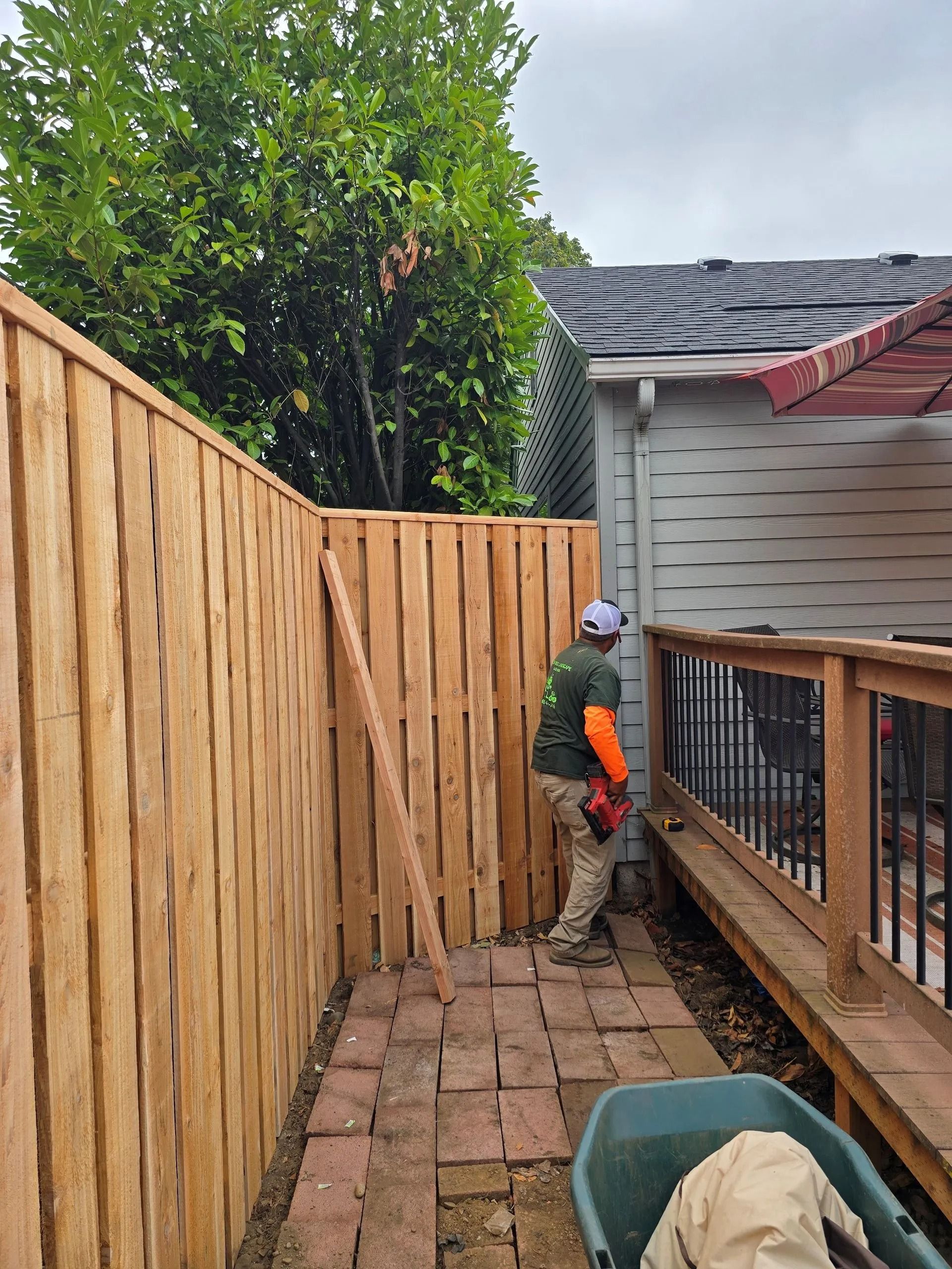 A person wearing a high-visibility orange sleeve works on a wooden fence next to a brick patio and a deck.