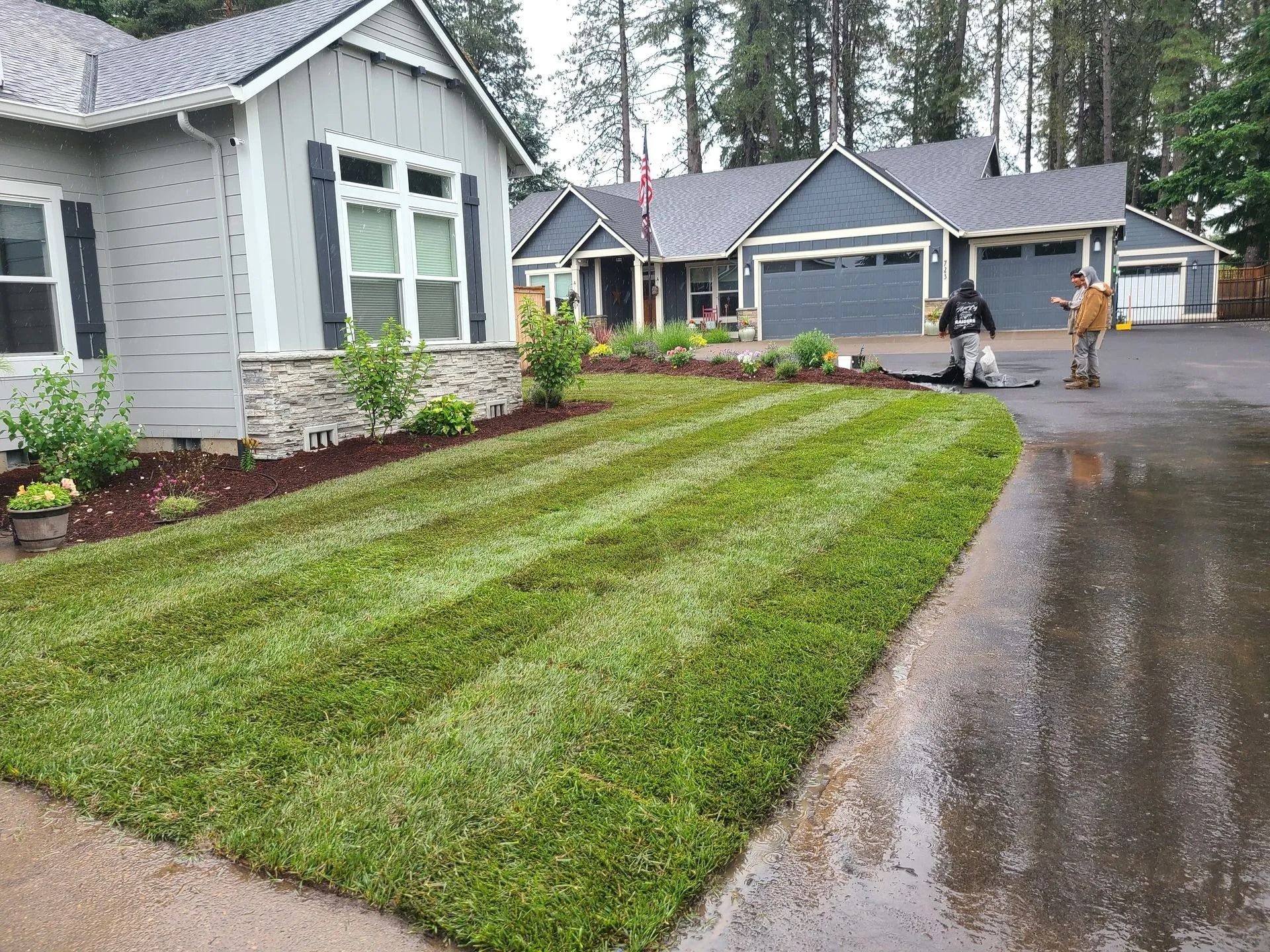 A freshly mown lawn in front of a gray house with a wet driveway and two people standing near garage doors.