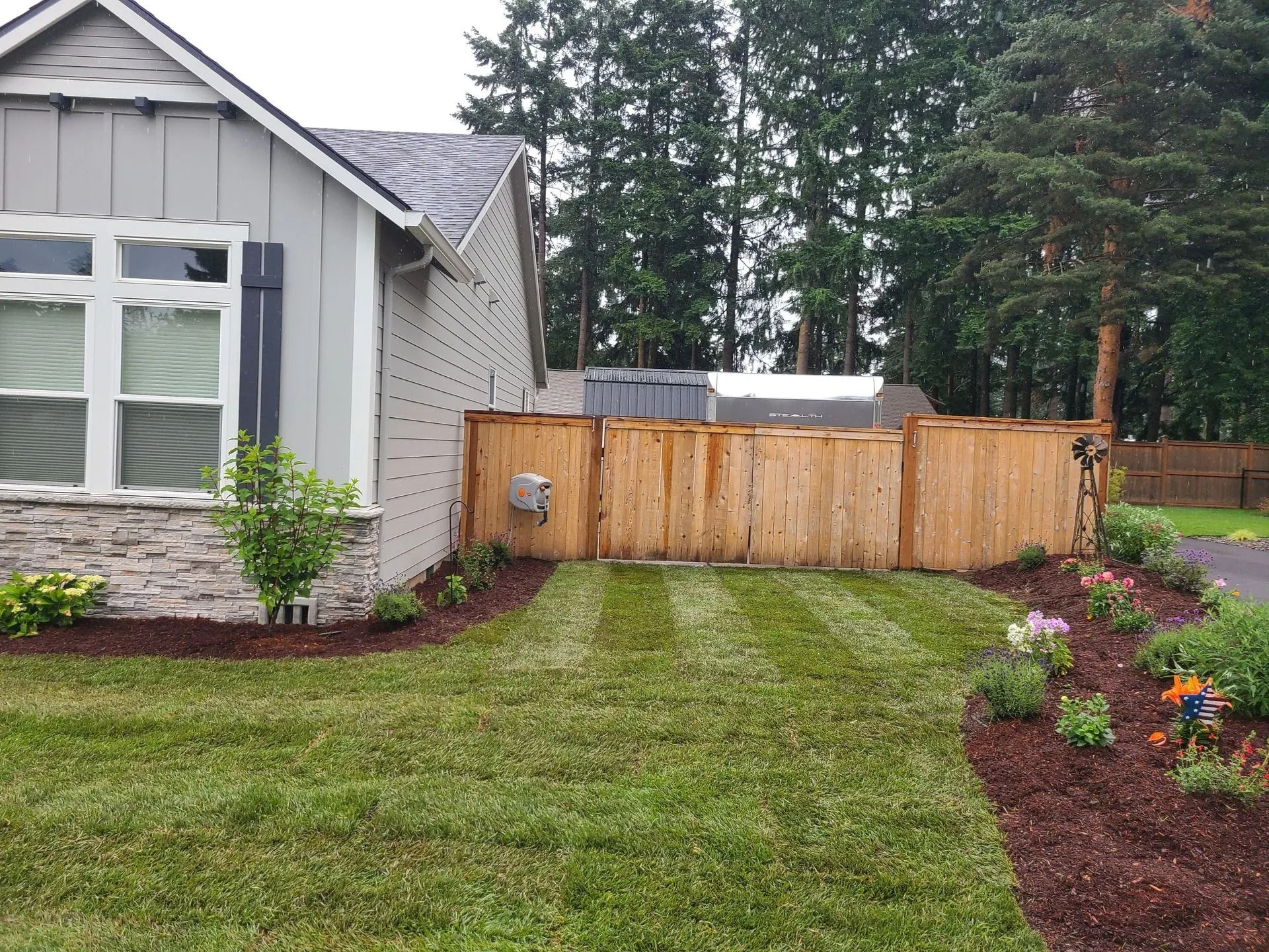 A side view of a gray house with a stone base, a manicured lawn, fresh mulch, and a new wood fence in a suburban backyard.