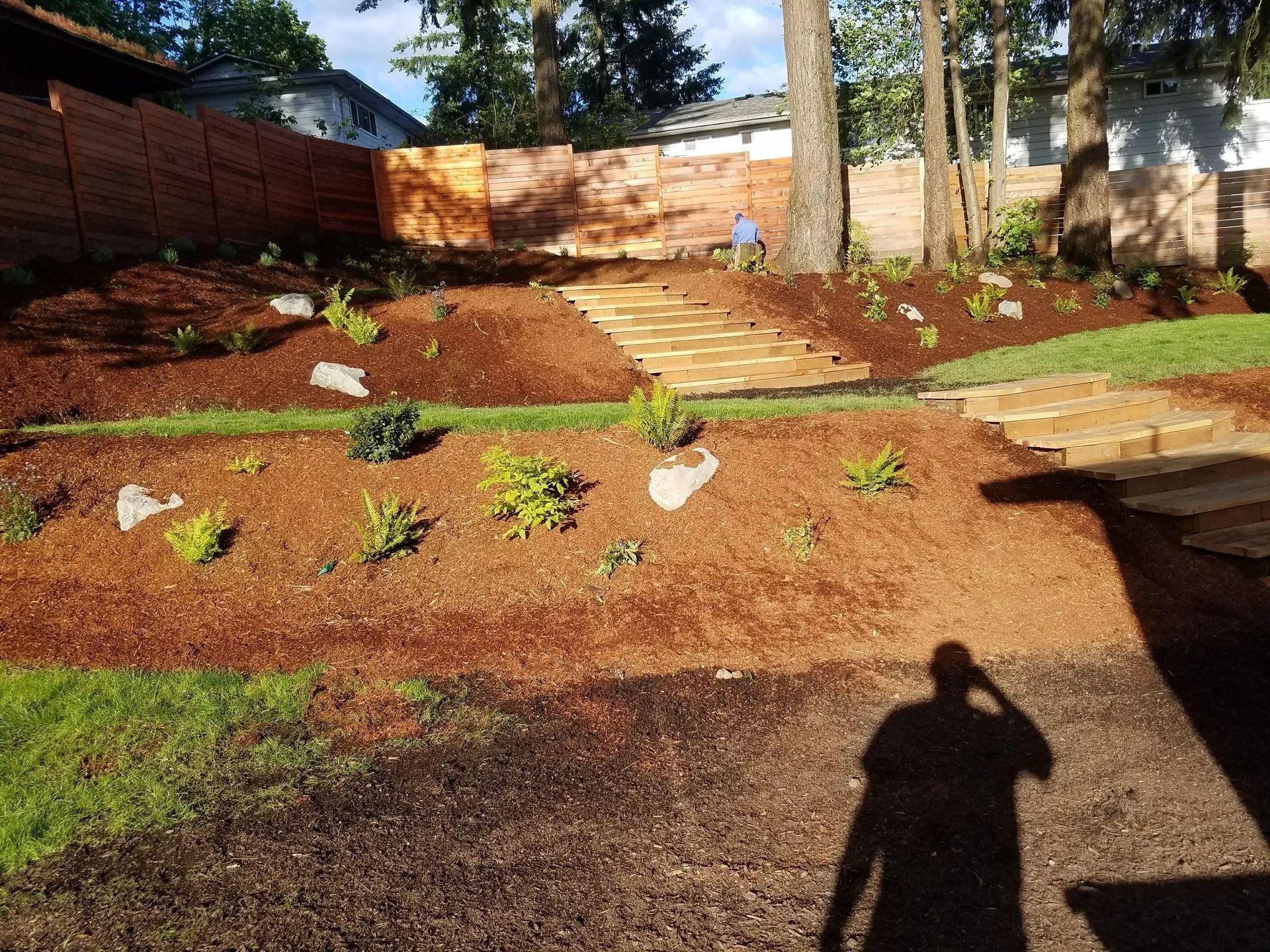 A landscaped backyard featuring tiered mulch garden beds, stone steps leading up a slope, and a wooden fence at the back.