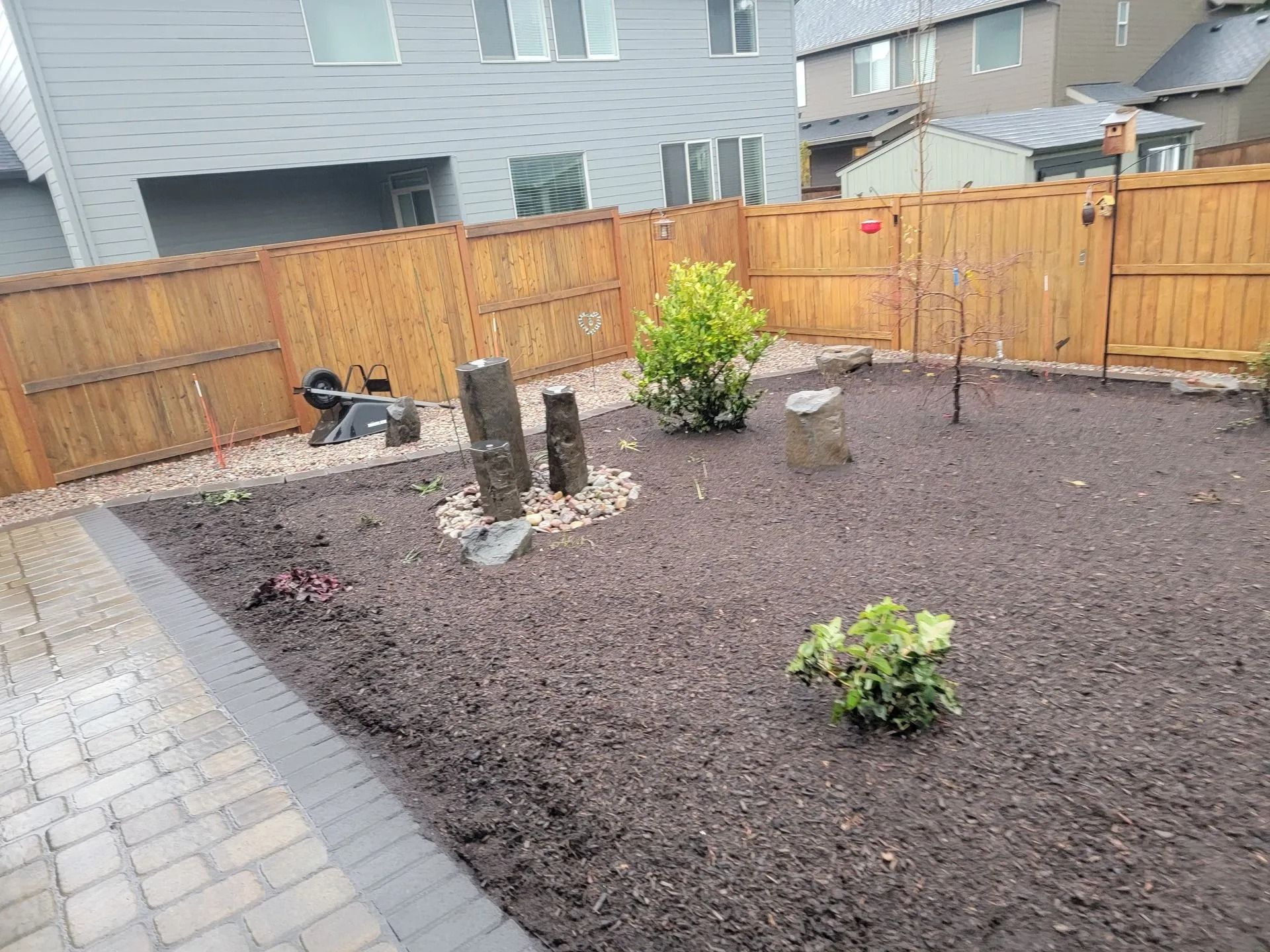 A backyard with a paver patio, a dark mulch area featuring two small shrubs, and a wooden privacy fence.