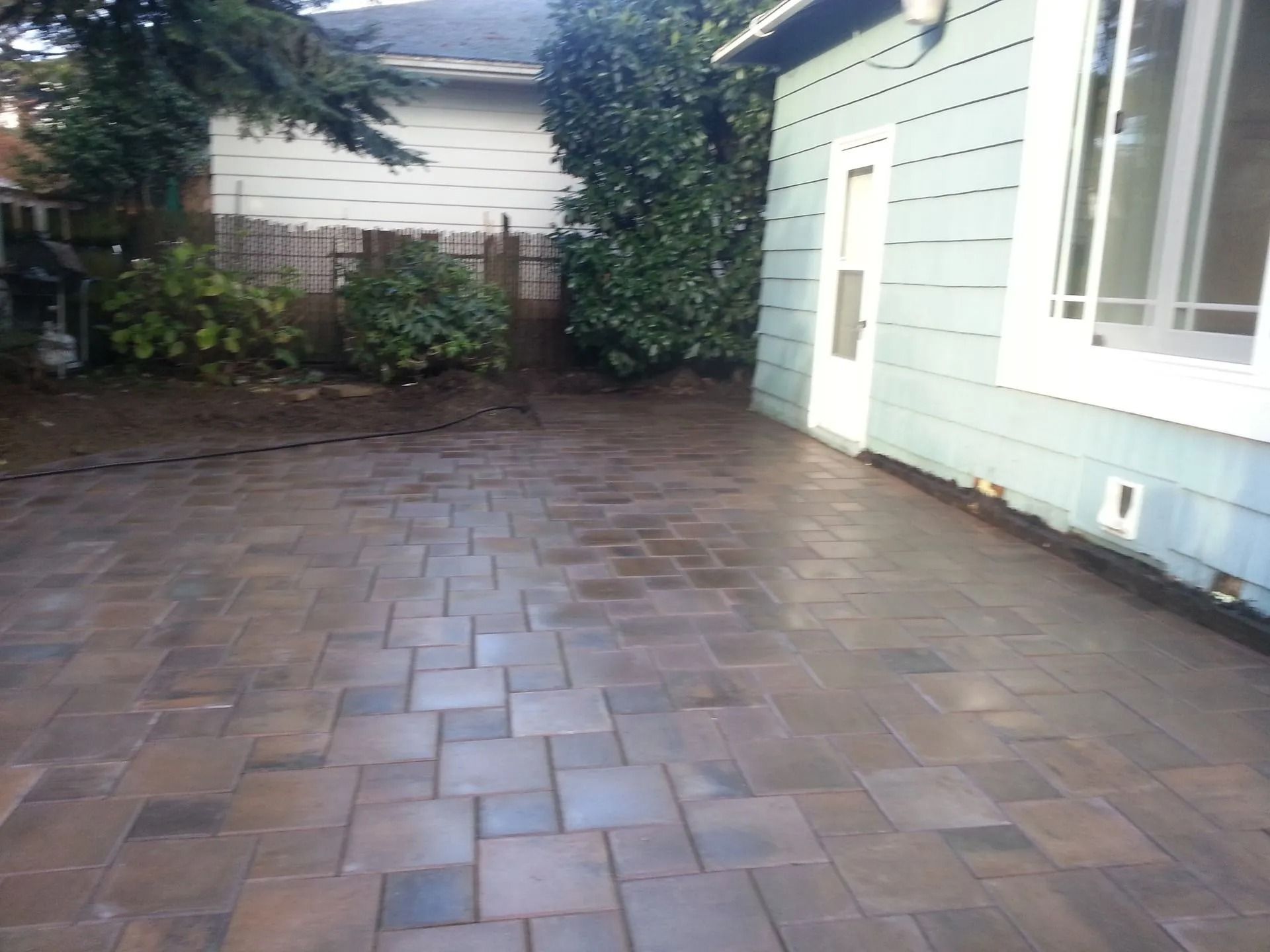 A new brown-toned paver patio beside a light blue house exterior with a door and window.