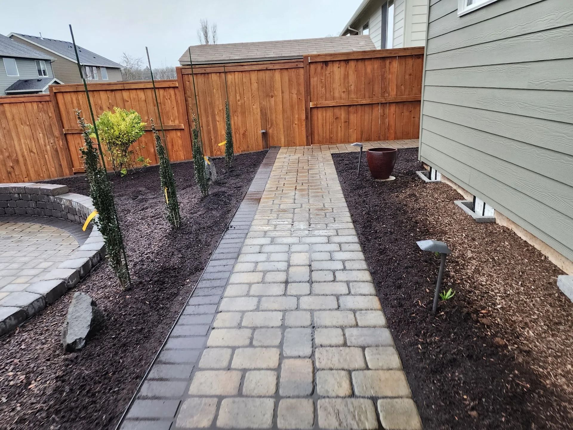 A paved stone walkway leads through a landscaped backyard with dark mulch, small green plants, and a wooden fence.