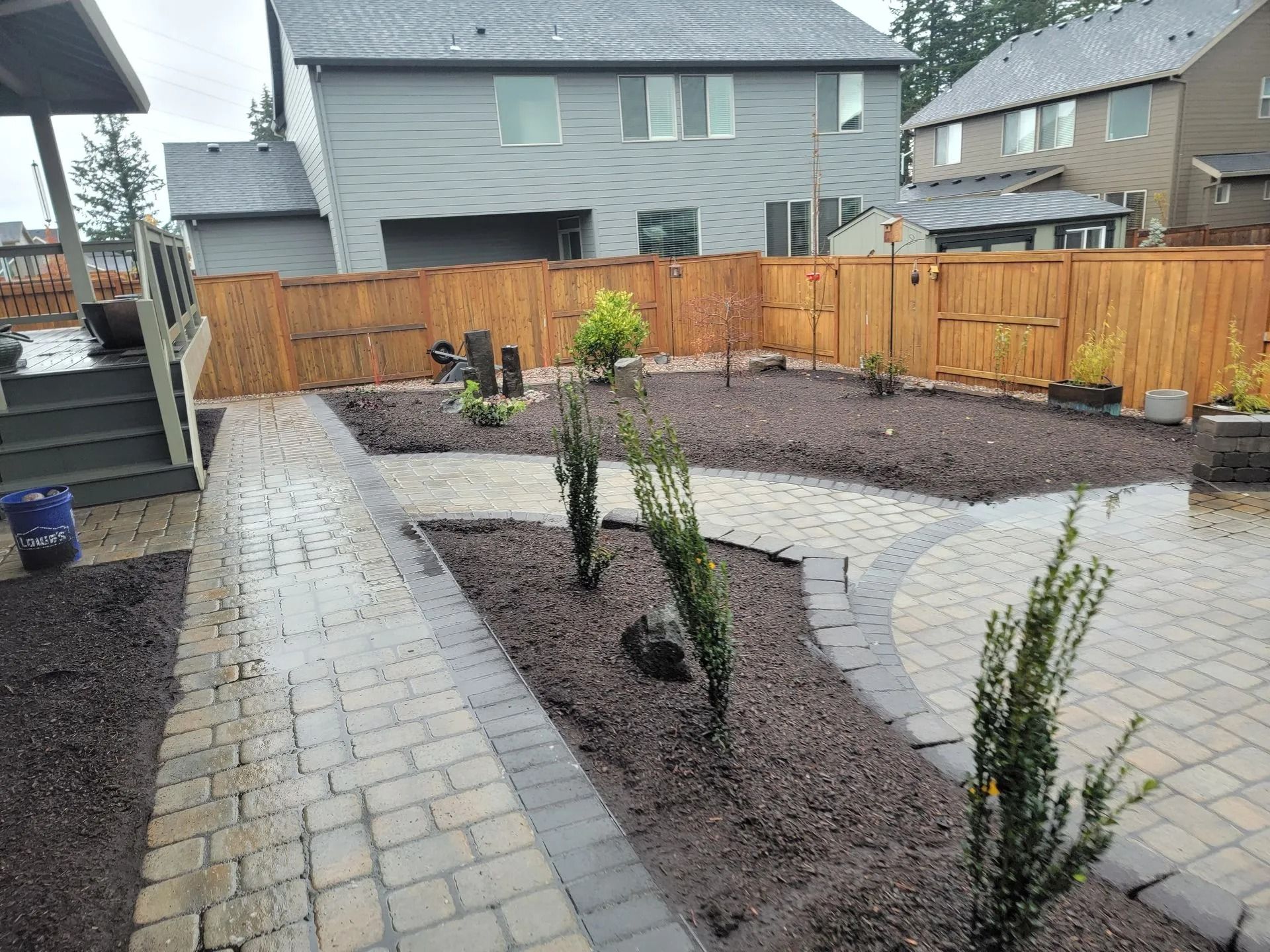 A backyard featuring a stone paver path and circular patio surrounded by dark mulch, young shrubs, and a wood fence.