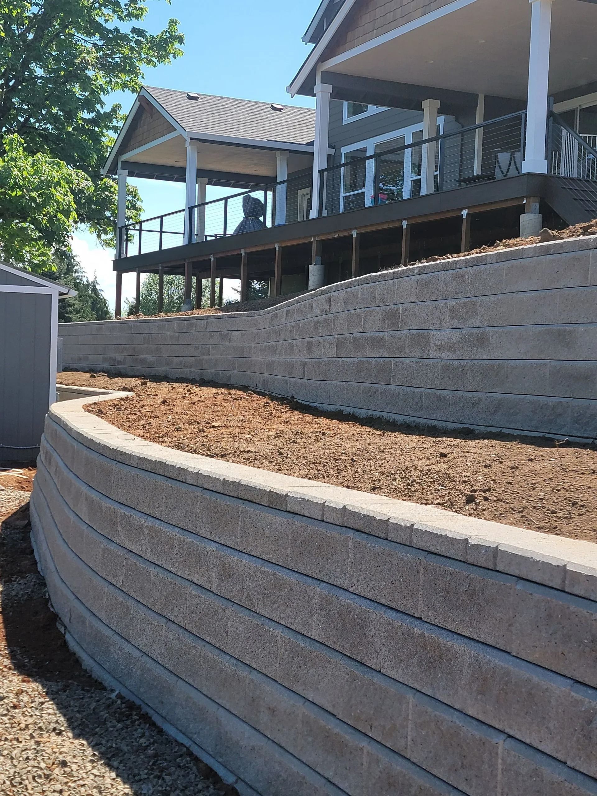 A terraced beige stone retaining wall holding back a mulched hillside below a house with a large wooden deck.
