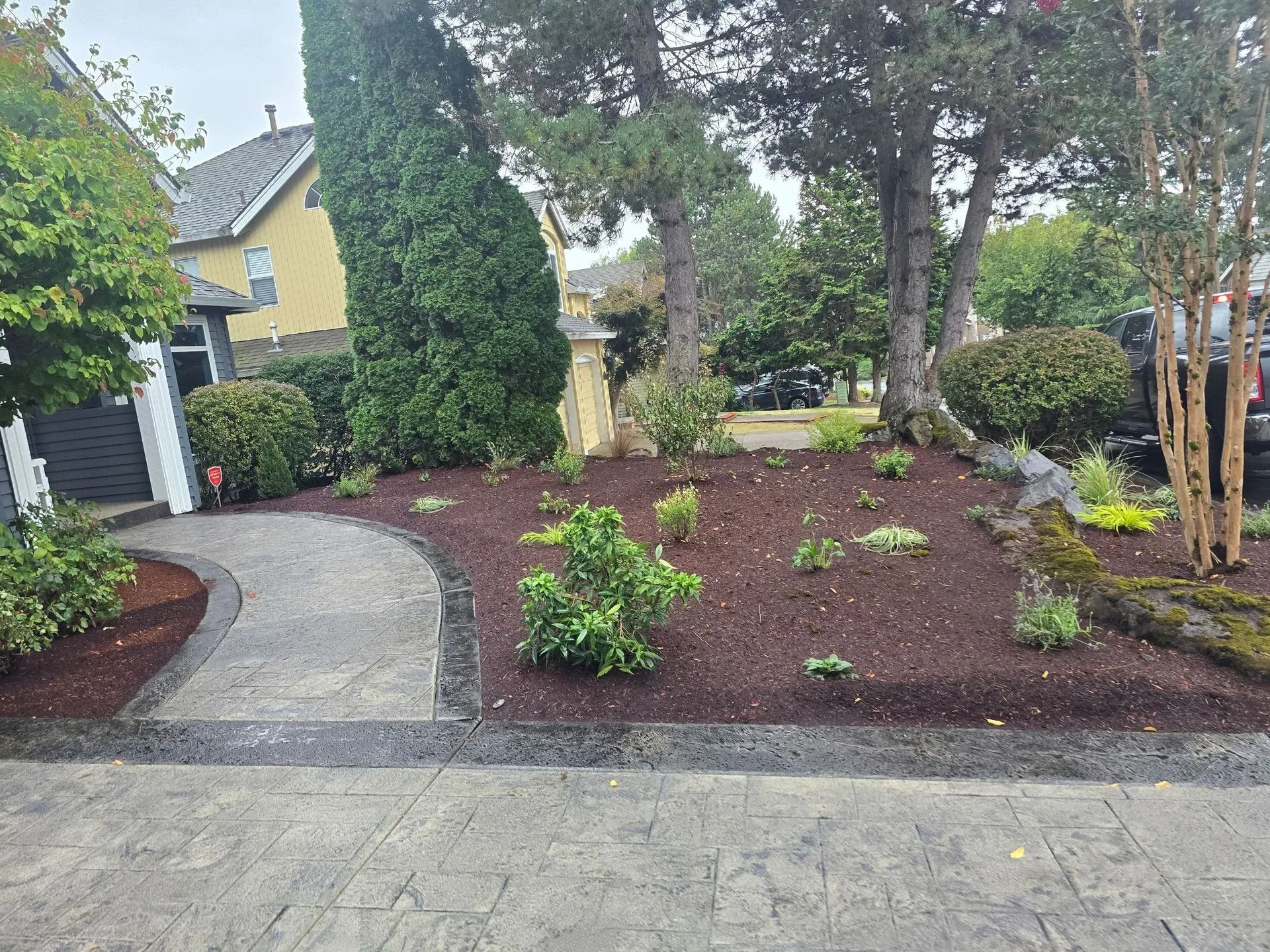 A paved walkway leads to a house entrance, past a landscaped yard with fresh dark mulch and small green shrubs.