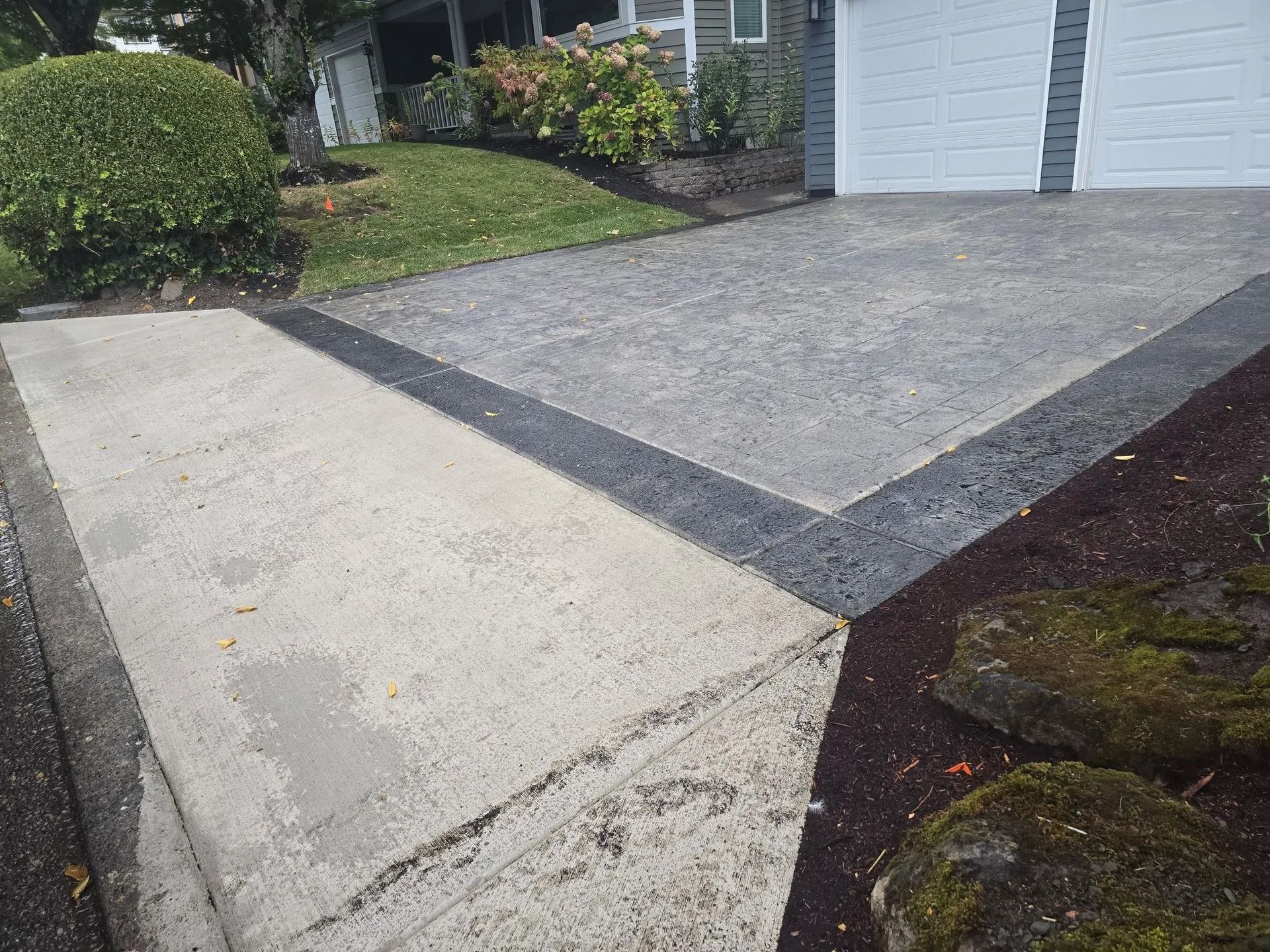 A driveway featuring a section of standard grey concrete and a section of textured, dark-bordered grey pavers.