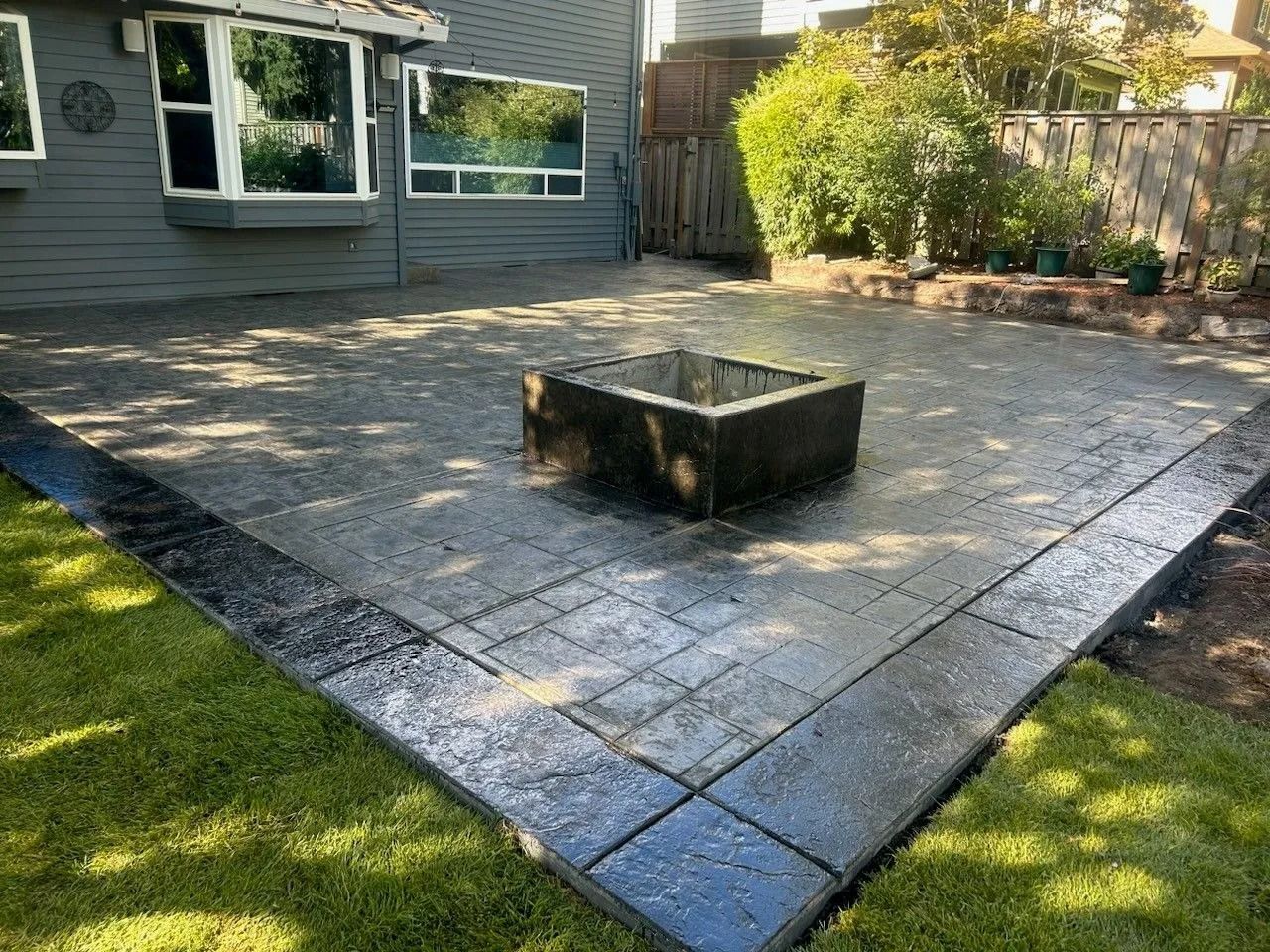 A backyard patio with gray textured pavers, a wide border, and a central square fire pit, adjacent to a gray house.