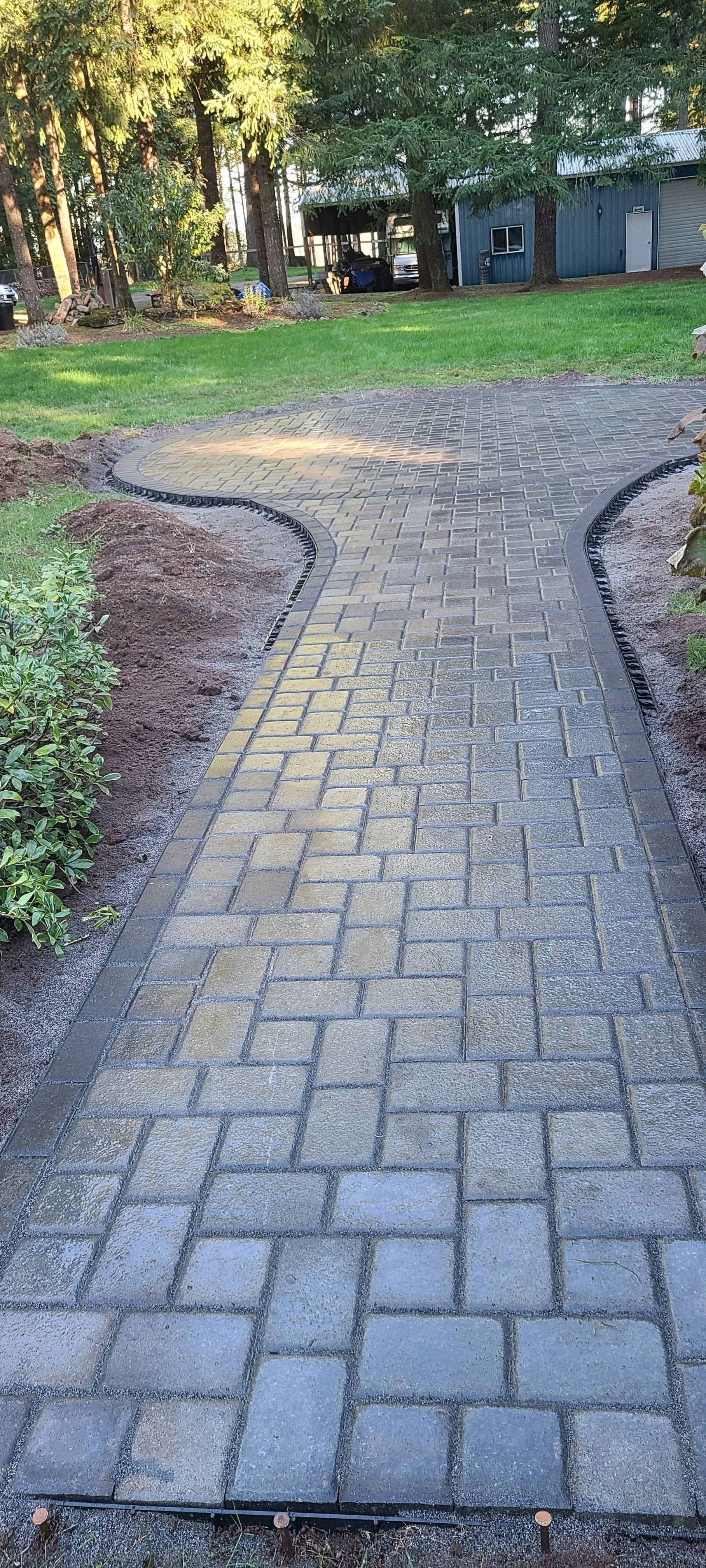 A stone paver walkway leads through a yard, flanked by mulch beds and green grass toward a house in the background.