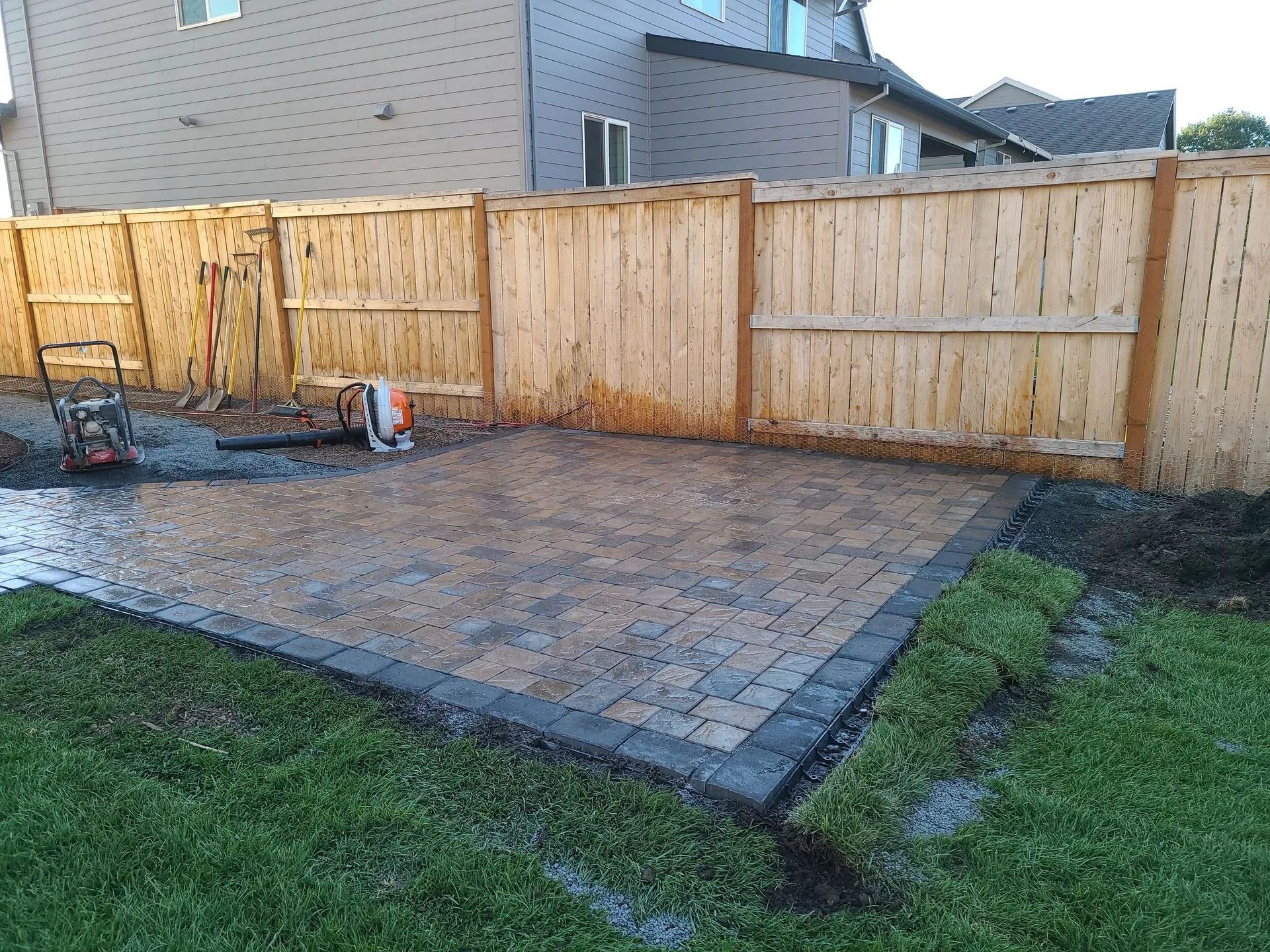 A newly installed paver patio in a backyard, bordered by a wooden fence, with a leaf blower nearby on the grass.