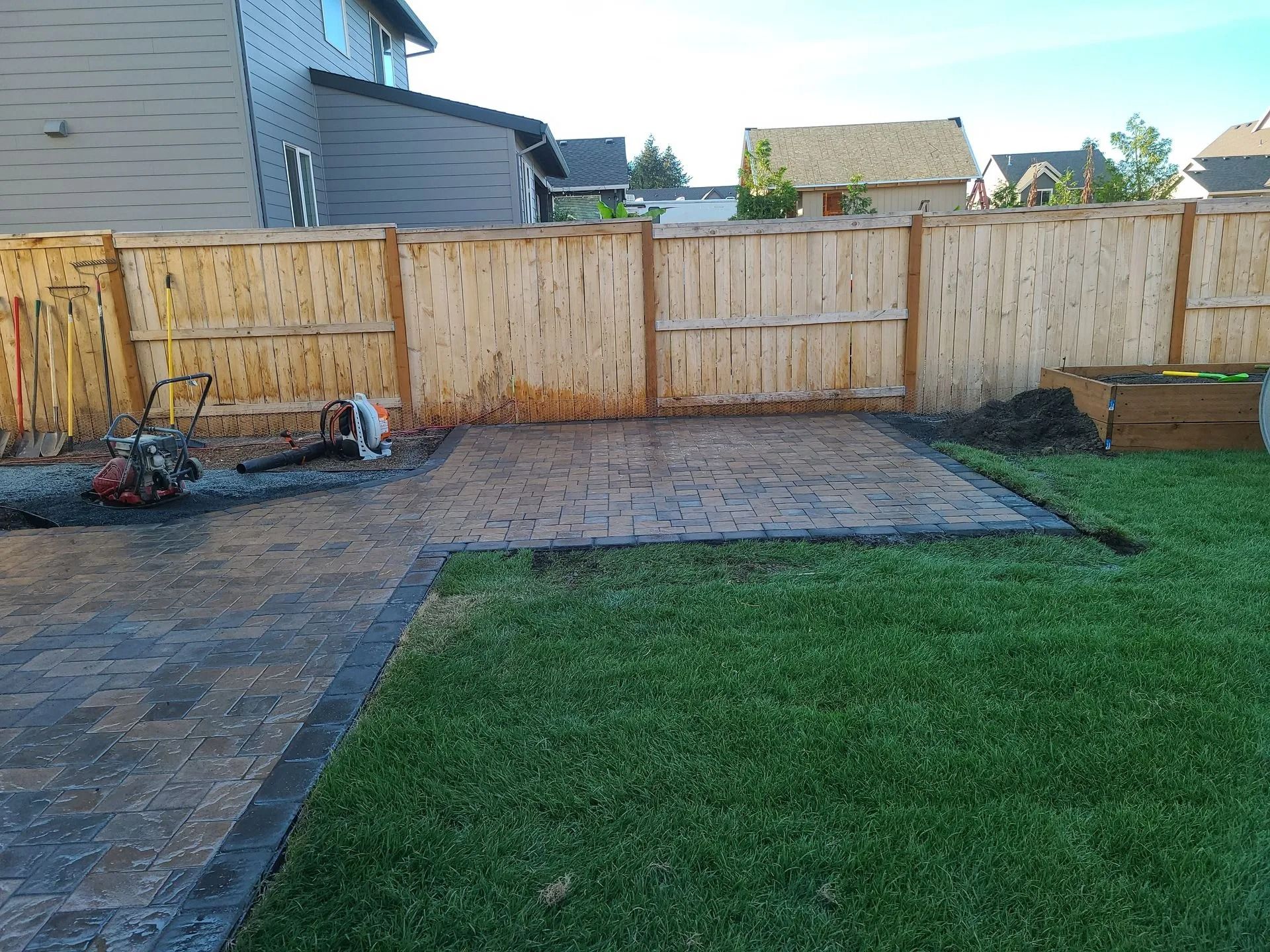 A newly installed paver patio area in a backyard, featuring a dark border and a wooden privacy fence in the background.