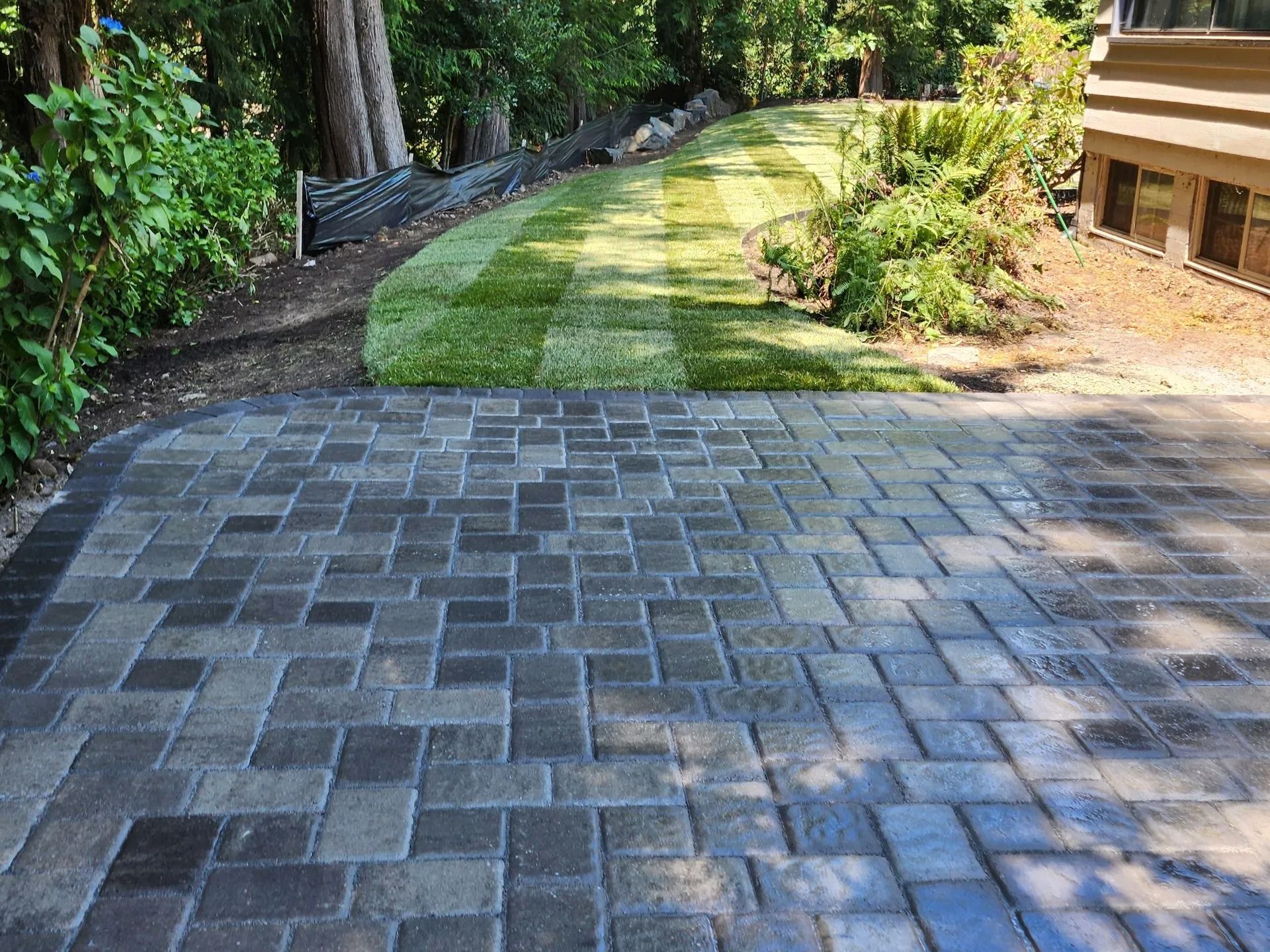 A stone paver patio abuts a freshly laid green grass lawn leading toward a house foundation and wooded area.