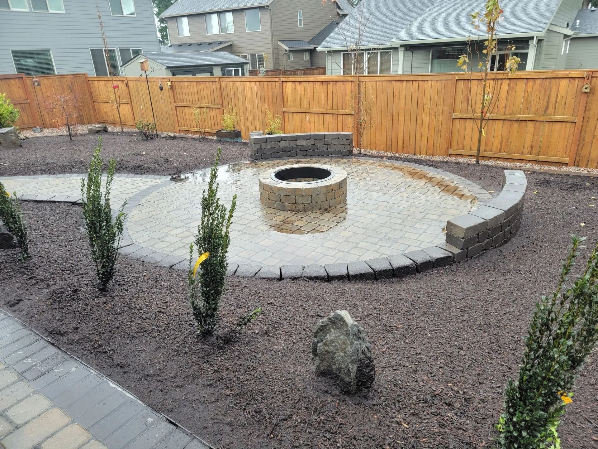 A round stone fire pit sits on a circular paved patio surrounded by dark mulch, landscaping, and a wooden privacy fence.