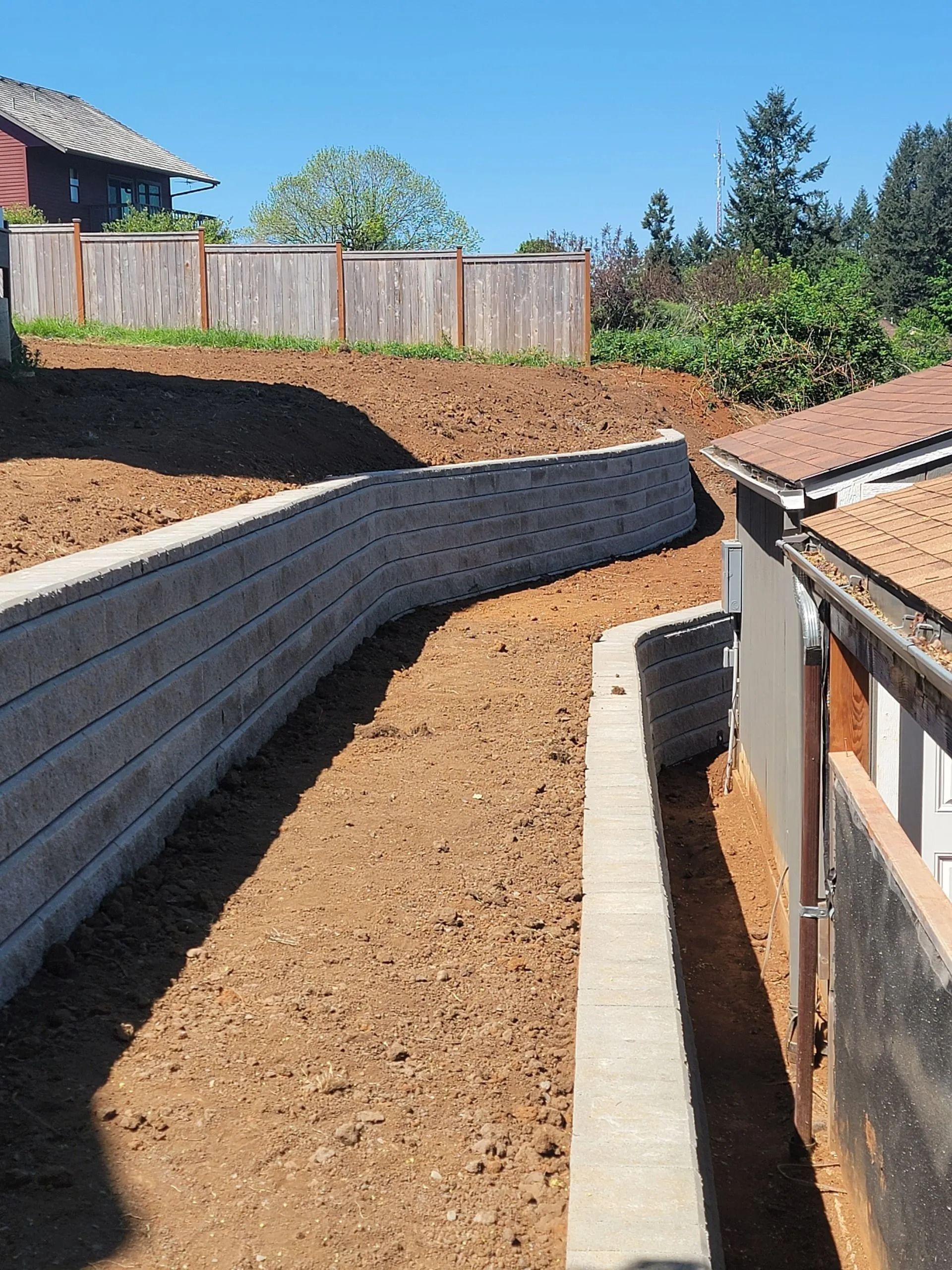 A backyard with two tan, tiered concrete block retaining walls and a dirt ground between them near a residential building.