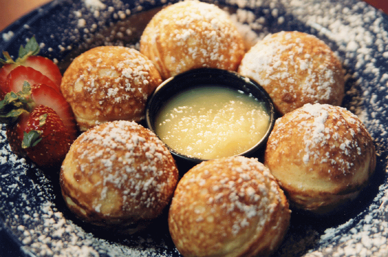 Poffertjes, powdered sugar, lemon curd dip, and sliced strawberries on a blue plate.