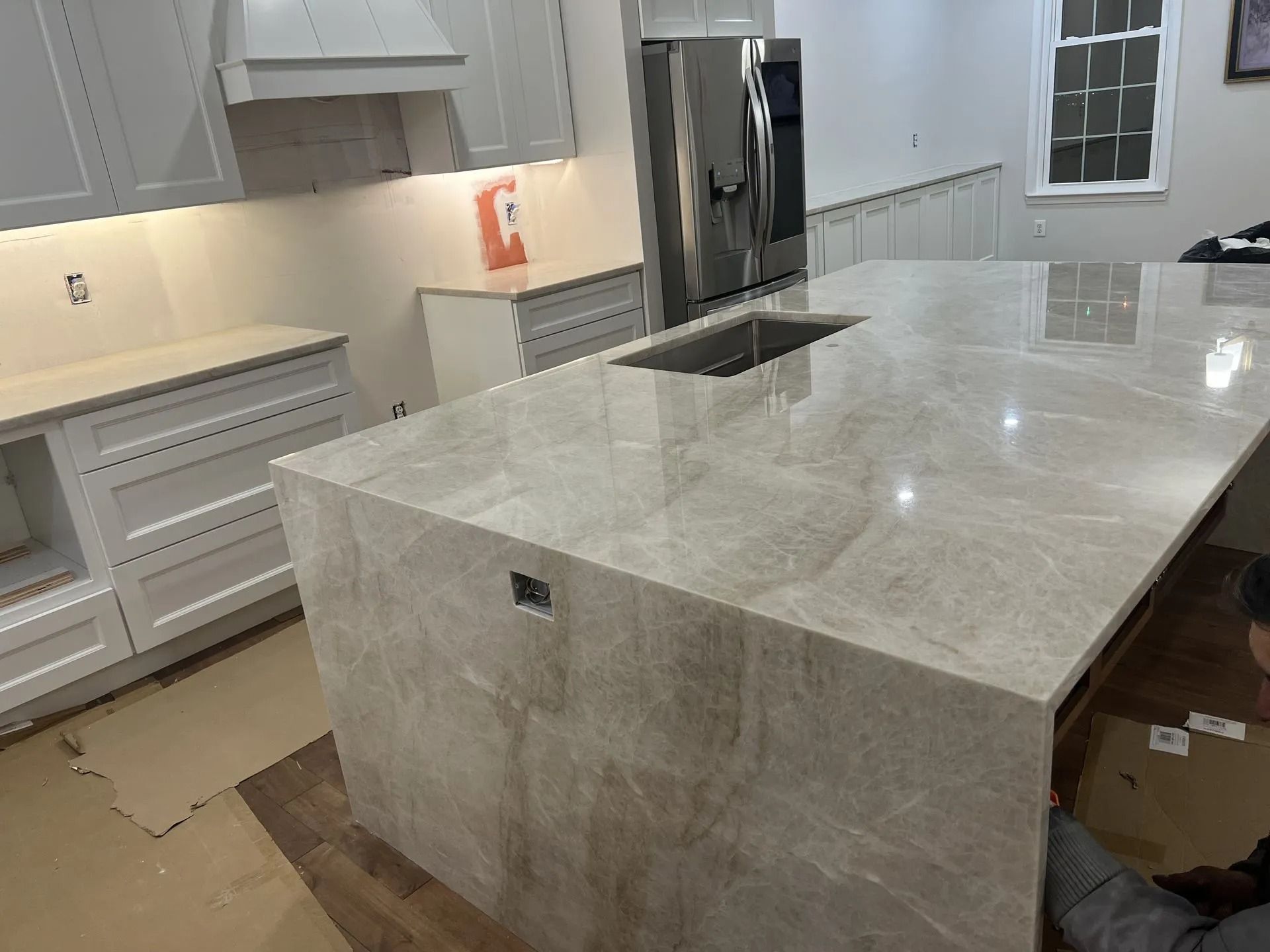 Modern kitchen with large marble island, white cabinets, stainless fridge, and unfinished floor area