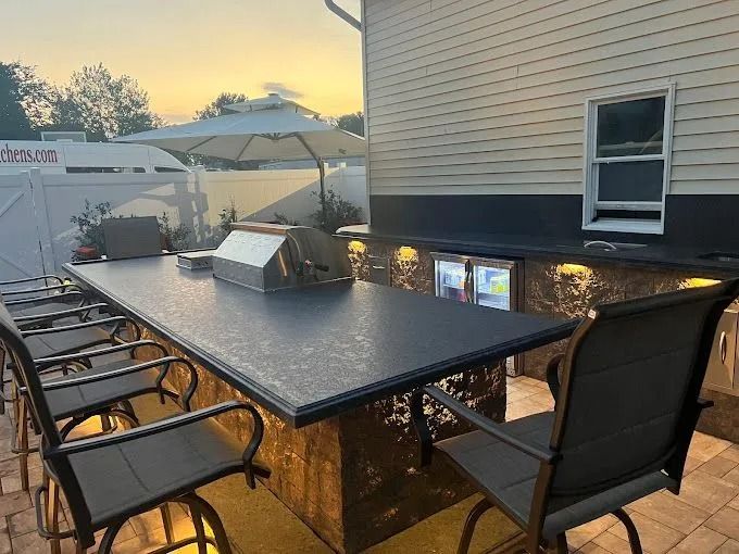 Outdoor patio dining area with black chairs, granite table, and built-in grill beside a house at sunset