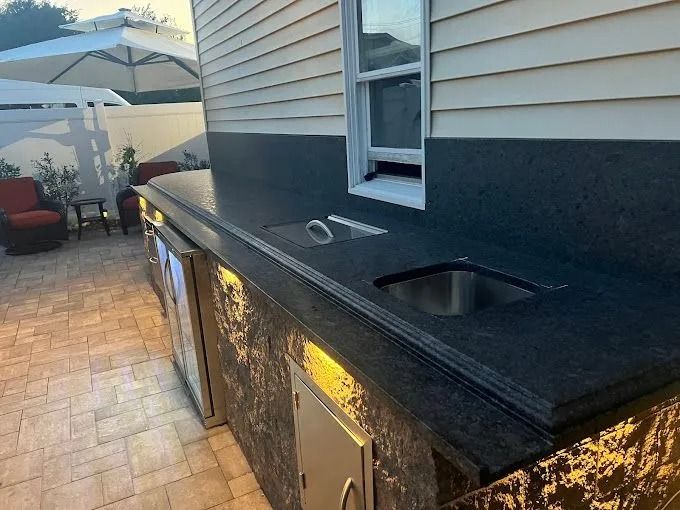 Outdoor patio with built-in grill and sink under a black countertop, beside a house wall