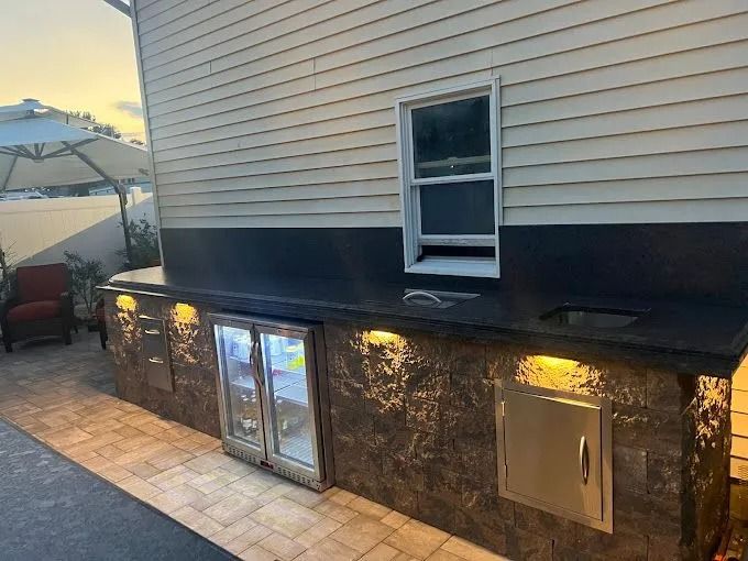 Basement entrance with double glass doors, lit stone wall, and utility access beneath beige siding at dusk