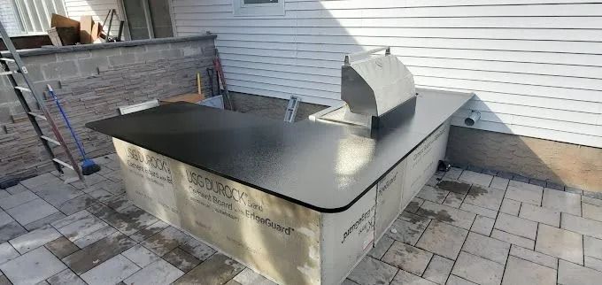 Outdoor stainless-steel grill with black cover on a stone patio beside a house