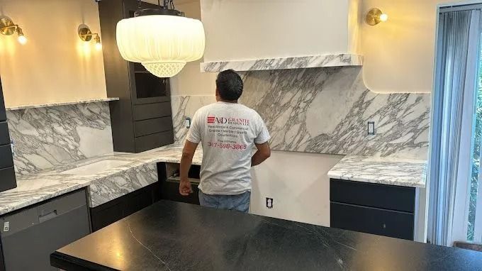 Person standing in a modern kitchen with marble counters, dark cabinets, and a white pendant light.