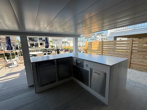 Outdoor kitchen with gray cabinets and countertop under a covered patio, beside a wooden fence.