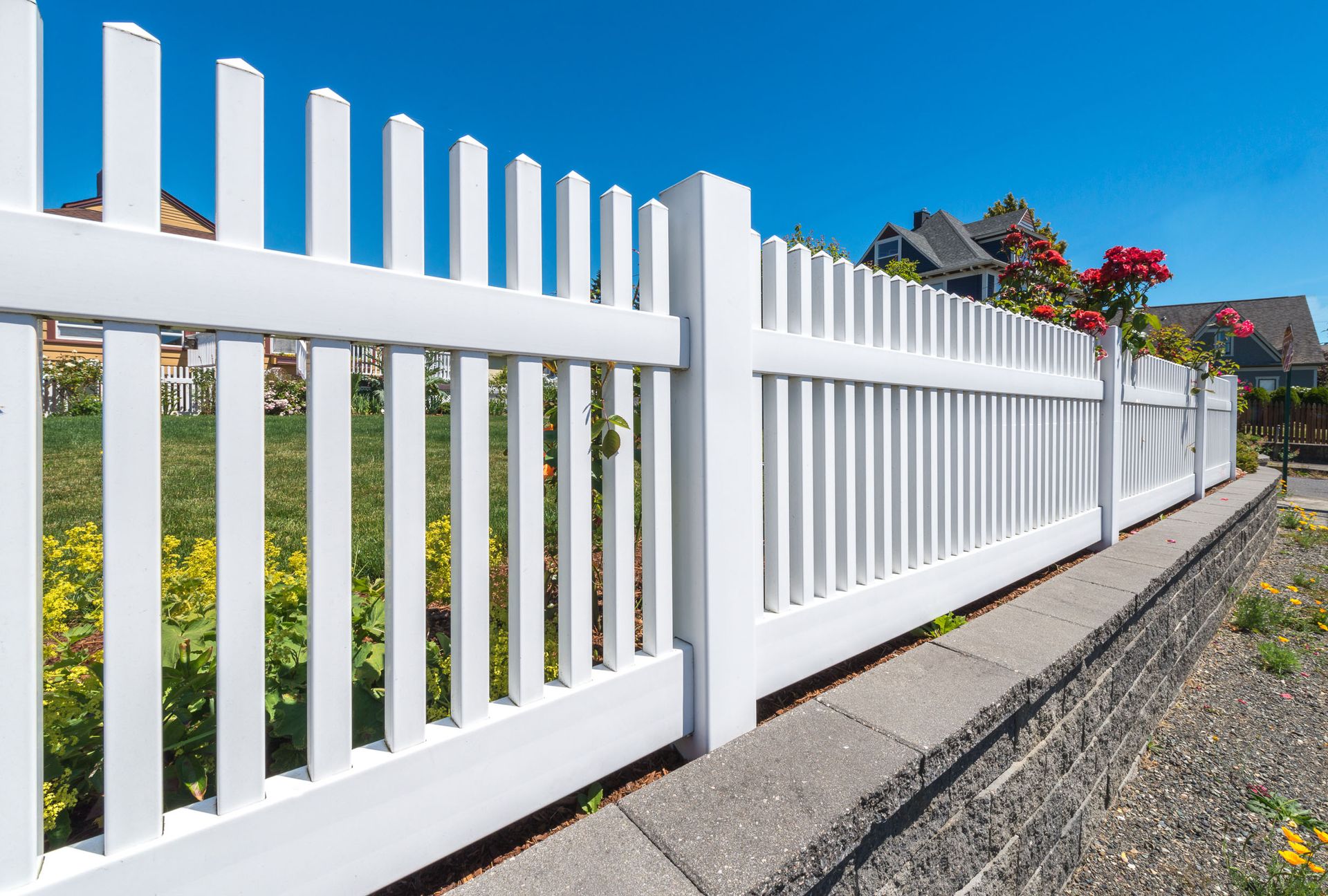 White picket fence along a roadside with blue sky and flowers in the background
