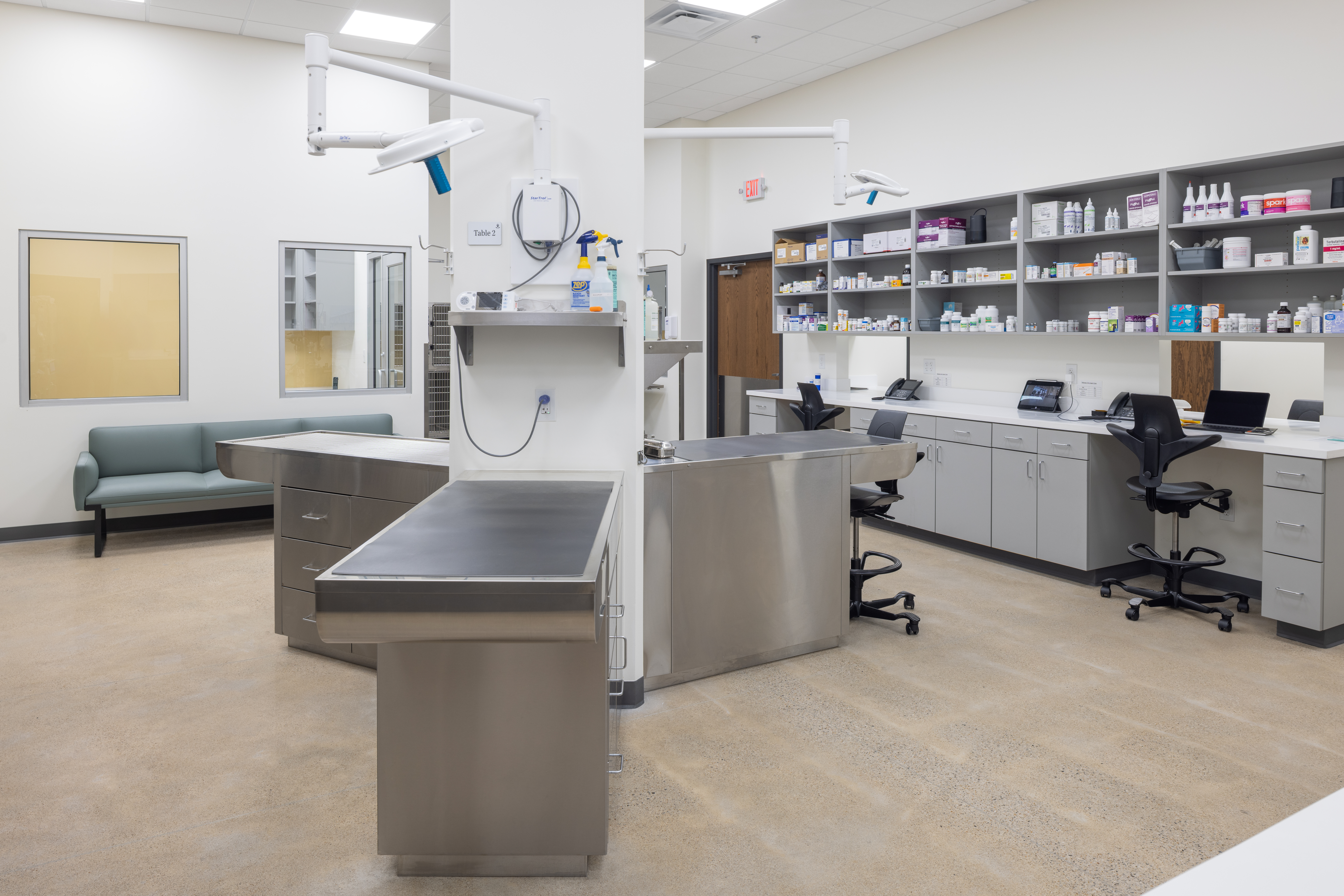 A modern veterinary examination room with stainless steel tables, medical supplies on shelves, and a gray seating bench.