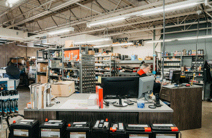 Inside an auto parts store. A person works at a counter behind car batteries, shelves with parts, and a computer.