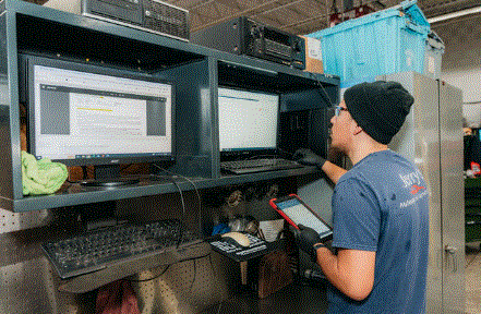 A man in a blue shirt and a beanie works at a computer station with two monitors, holding a tablet in a workshop setting.