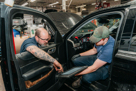 Two mechanics in a car interior, one with a tablet, working on the dashboard, in a garage.