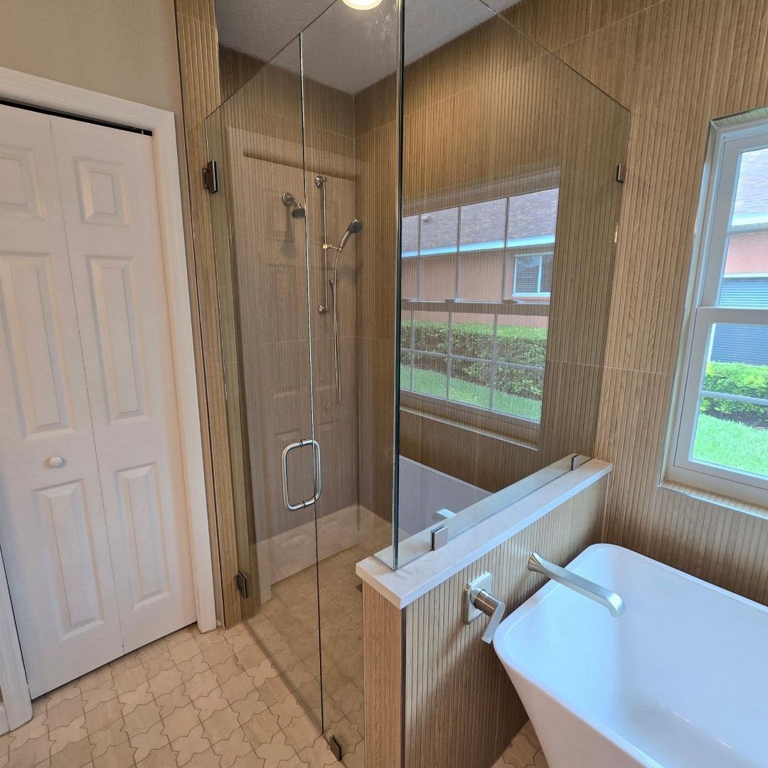 Accessible bathroom with glass shower, white tub, and wood-paneled walls beside a window