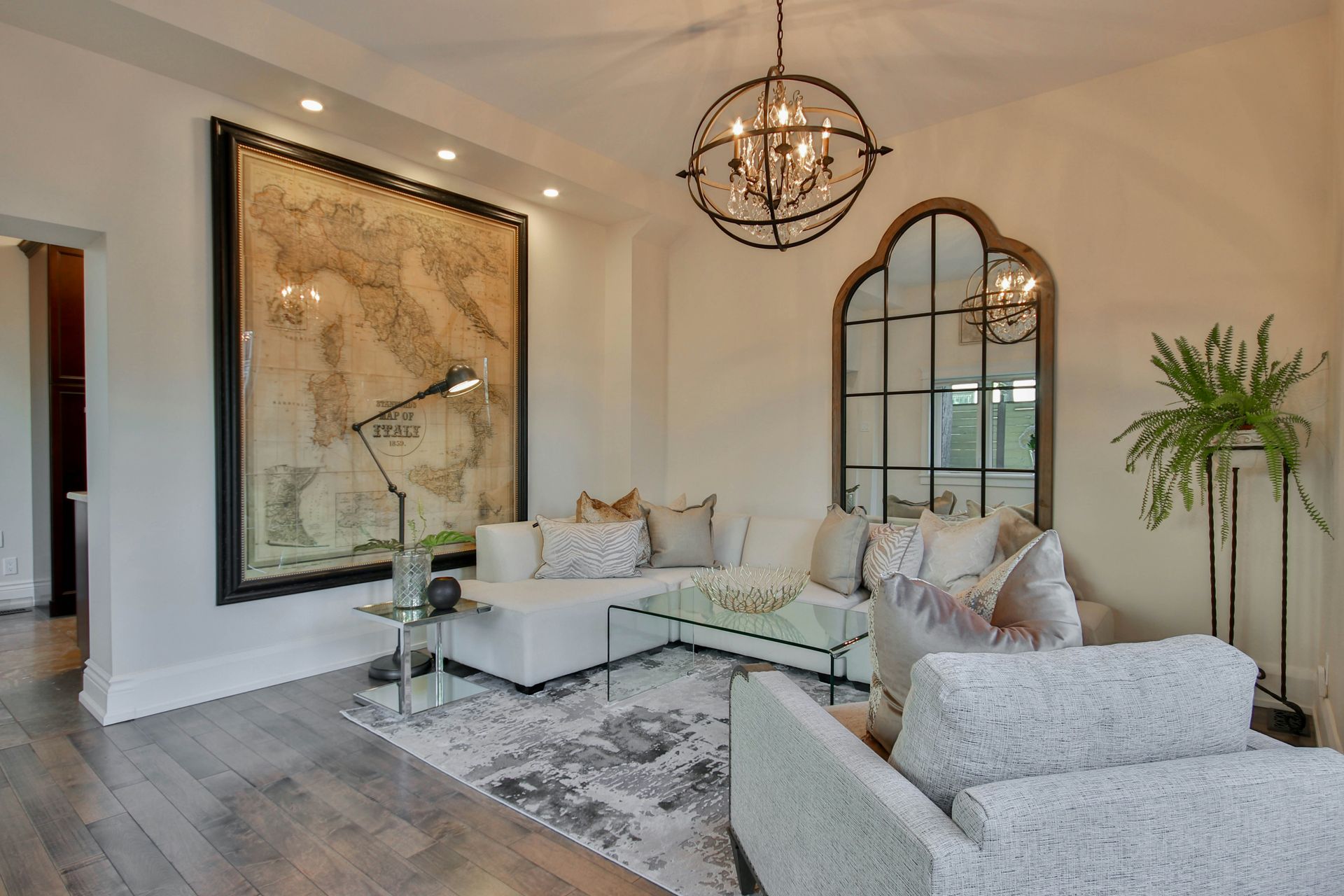 A bright, modern living room with a white sectional, large map artwork, a round chandelier, and an arched wall mirror.