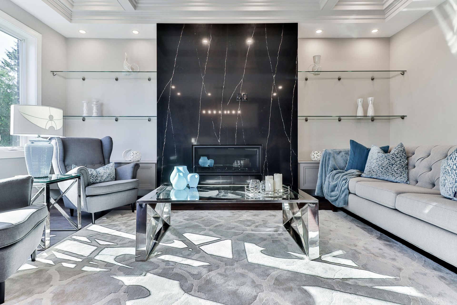 A modern living room with a black marble fireplace, gray armchairs, a tufted sofa, and glass shelves.