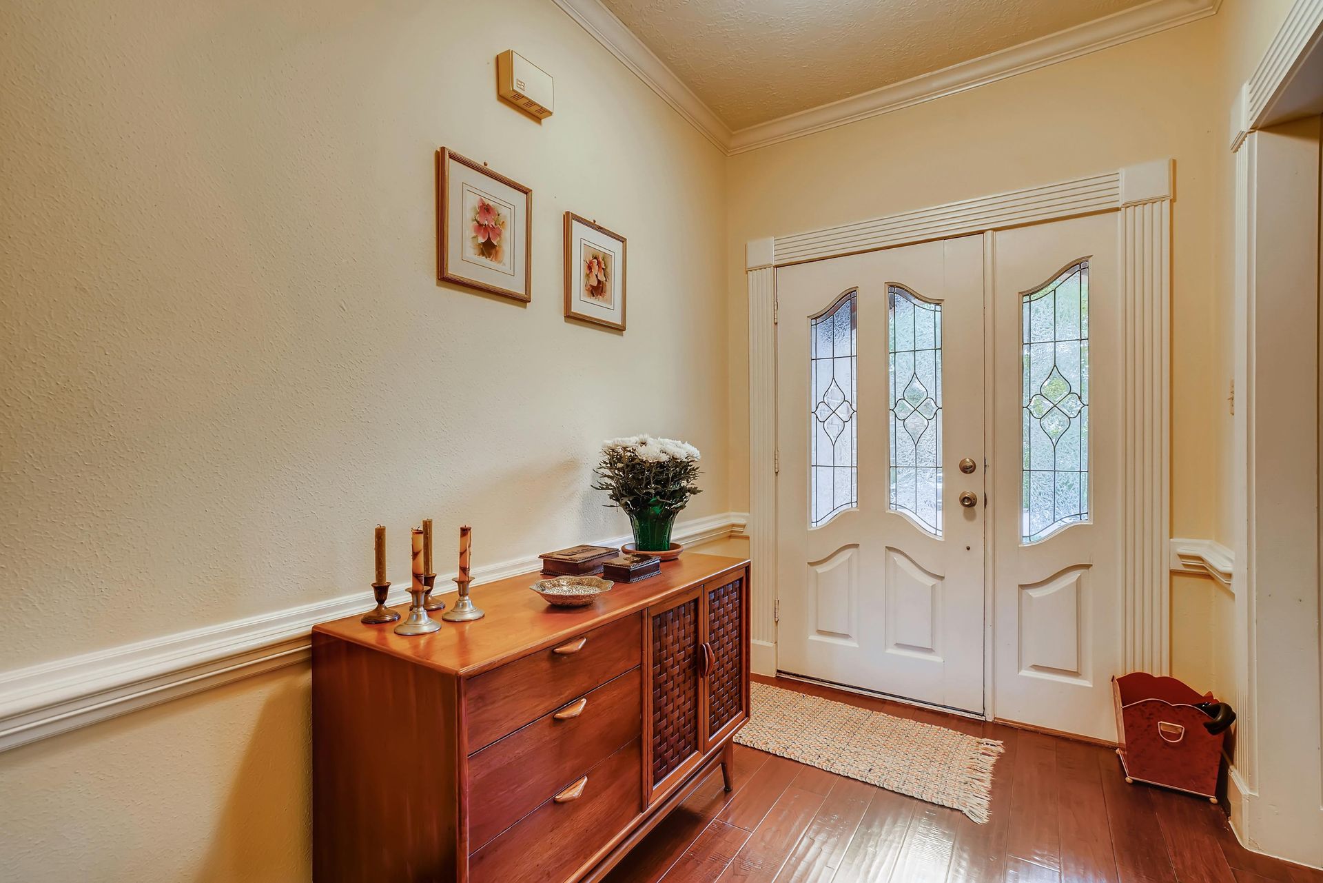 An entryway with a wooden dresser, floral arrangement, and framed wall art next to a white double front door.