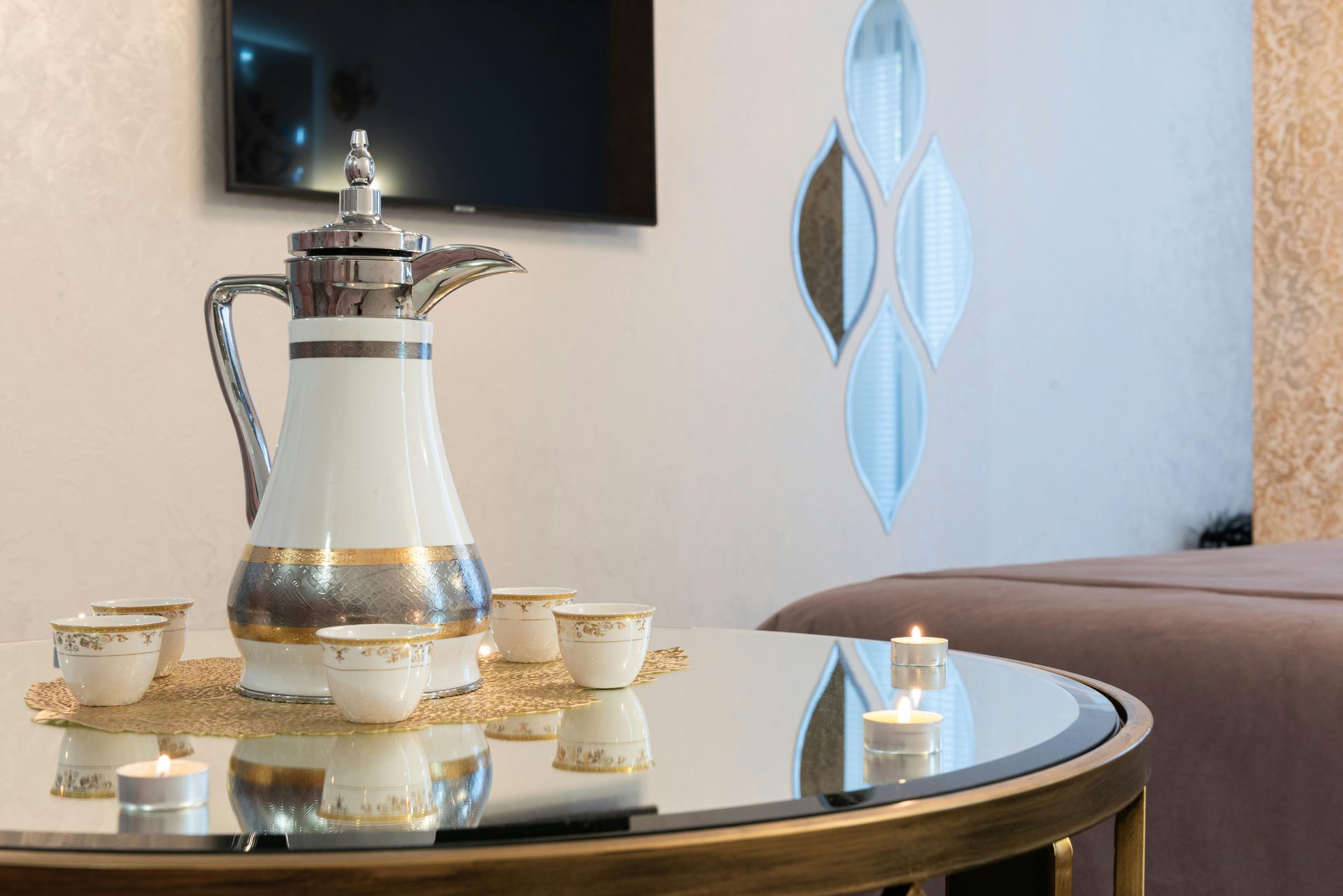 A traditional silver and white Arabic coffee pot sits on a glass table with small tea cups and lit candles in a room.