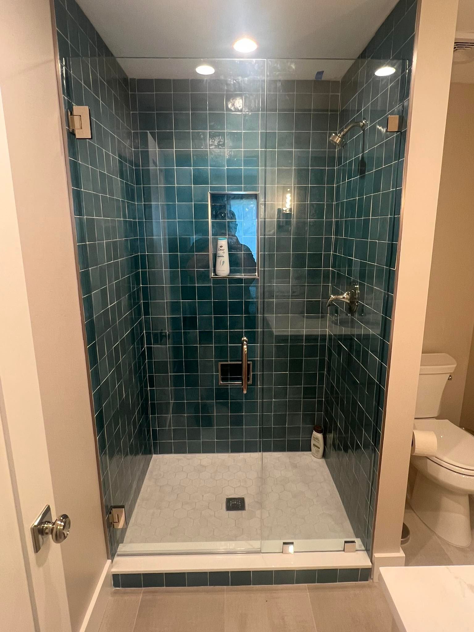 Walk-in shower with dark teal tile walls, glass door, and light stone floor under recessed lights