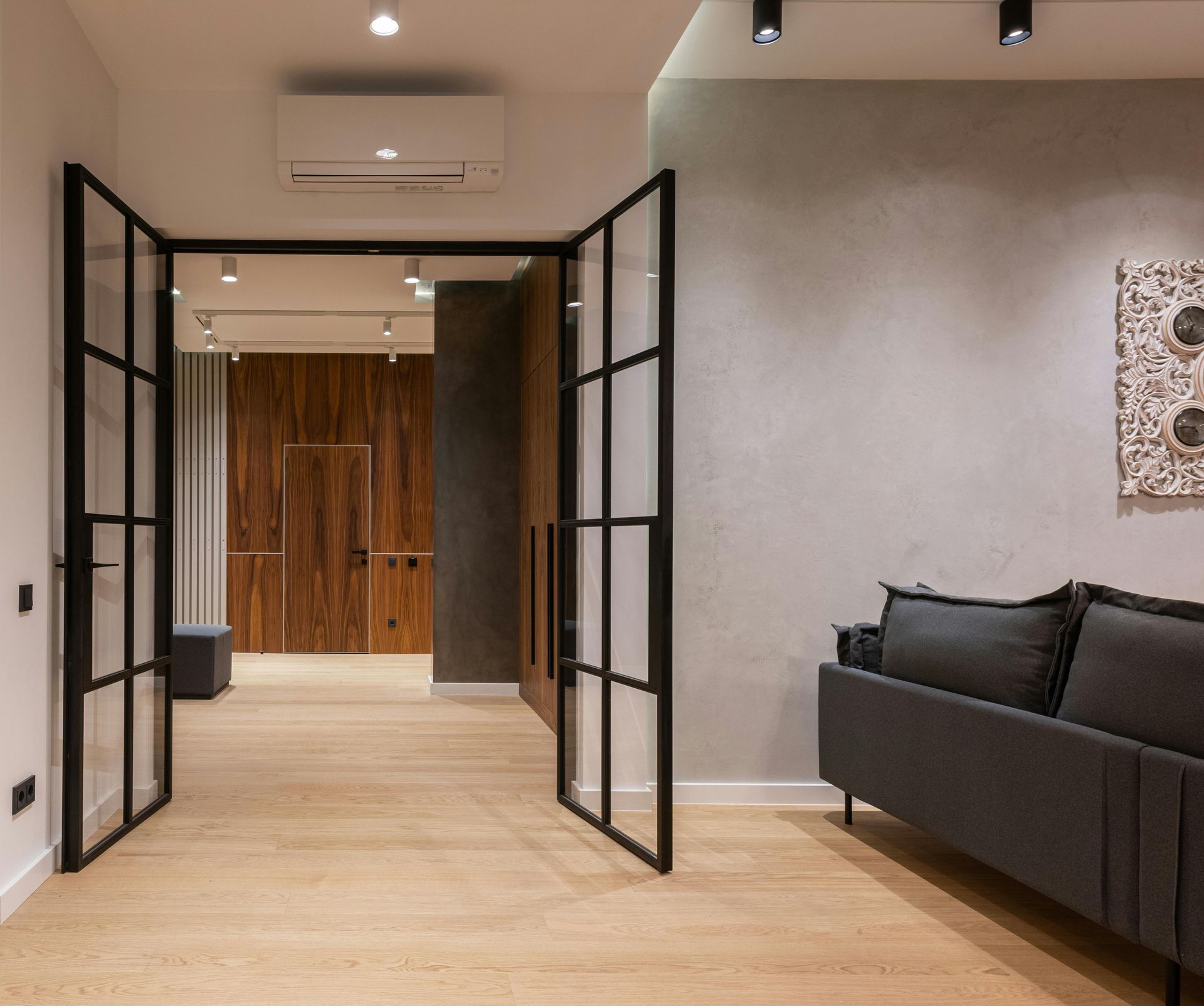 A modern interior view featuring black-framed glass double doors leading to a hallway, beside a grey sofa on light flooring.