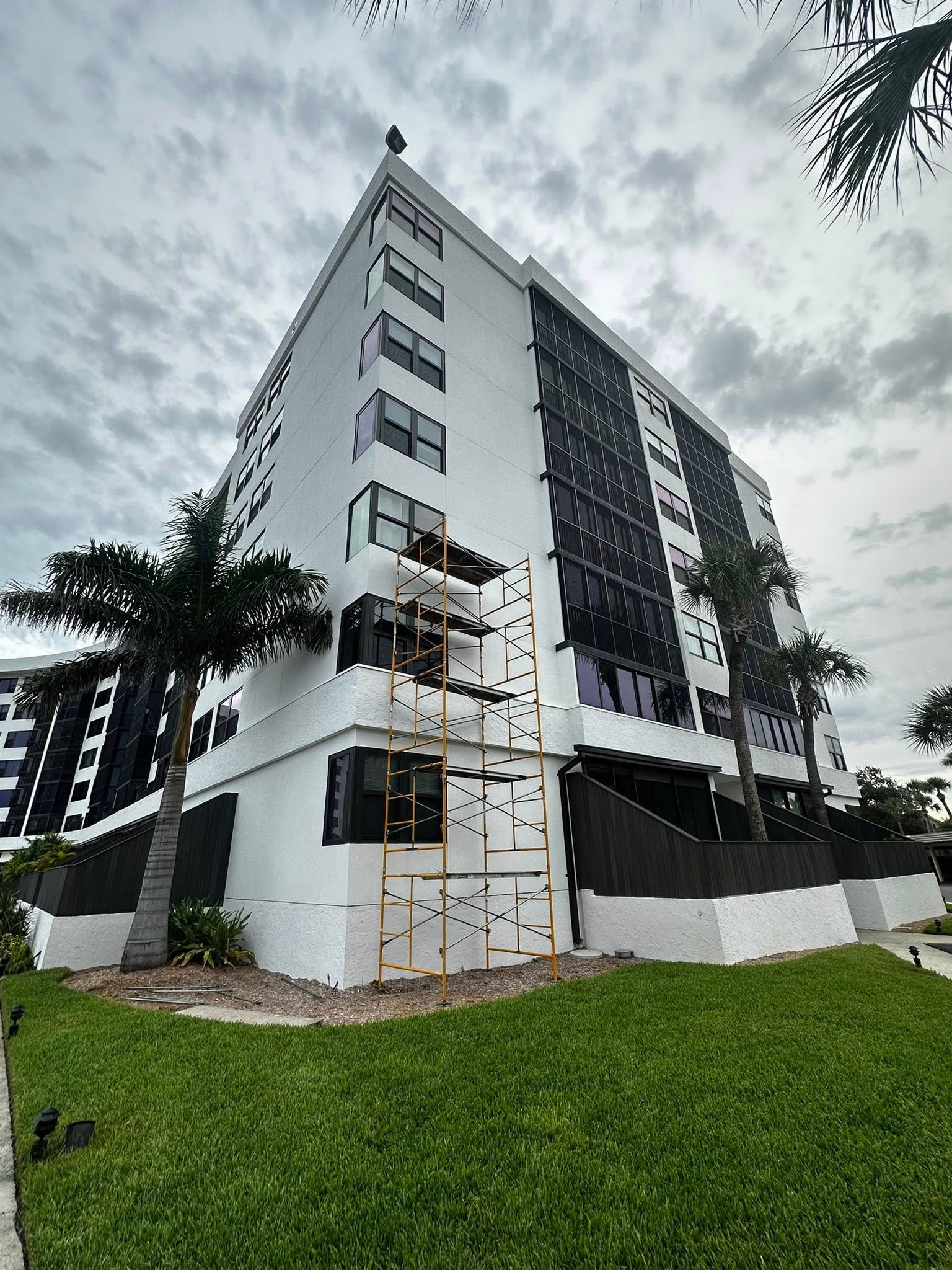 Low-angle view of a white apartment building with dark panels, scaffolding, and a green lawn under cloudy sky