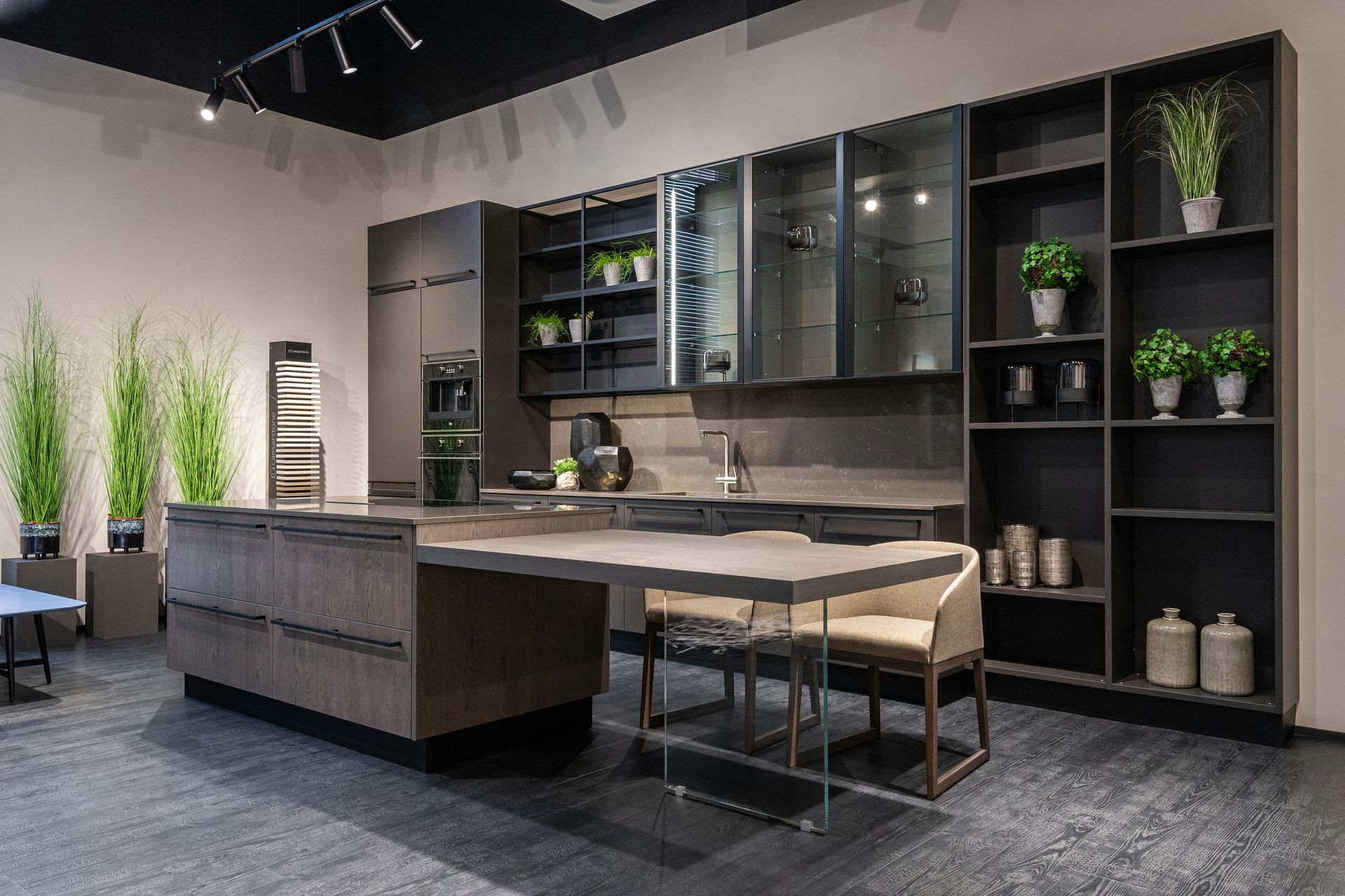 A modern, dark-toned kitchen display featuring a wood-grain island with a glass support, glass-front cabinets, and shelving.