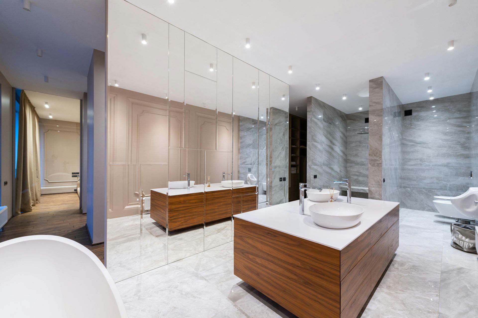 A spacious, modern bathroom with a wood-toned island vanity, marble floors, a large wall mirror, and gray tiled walls.