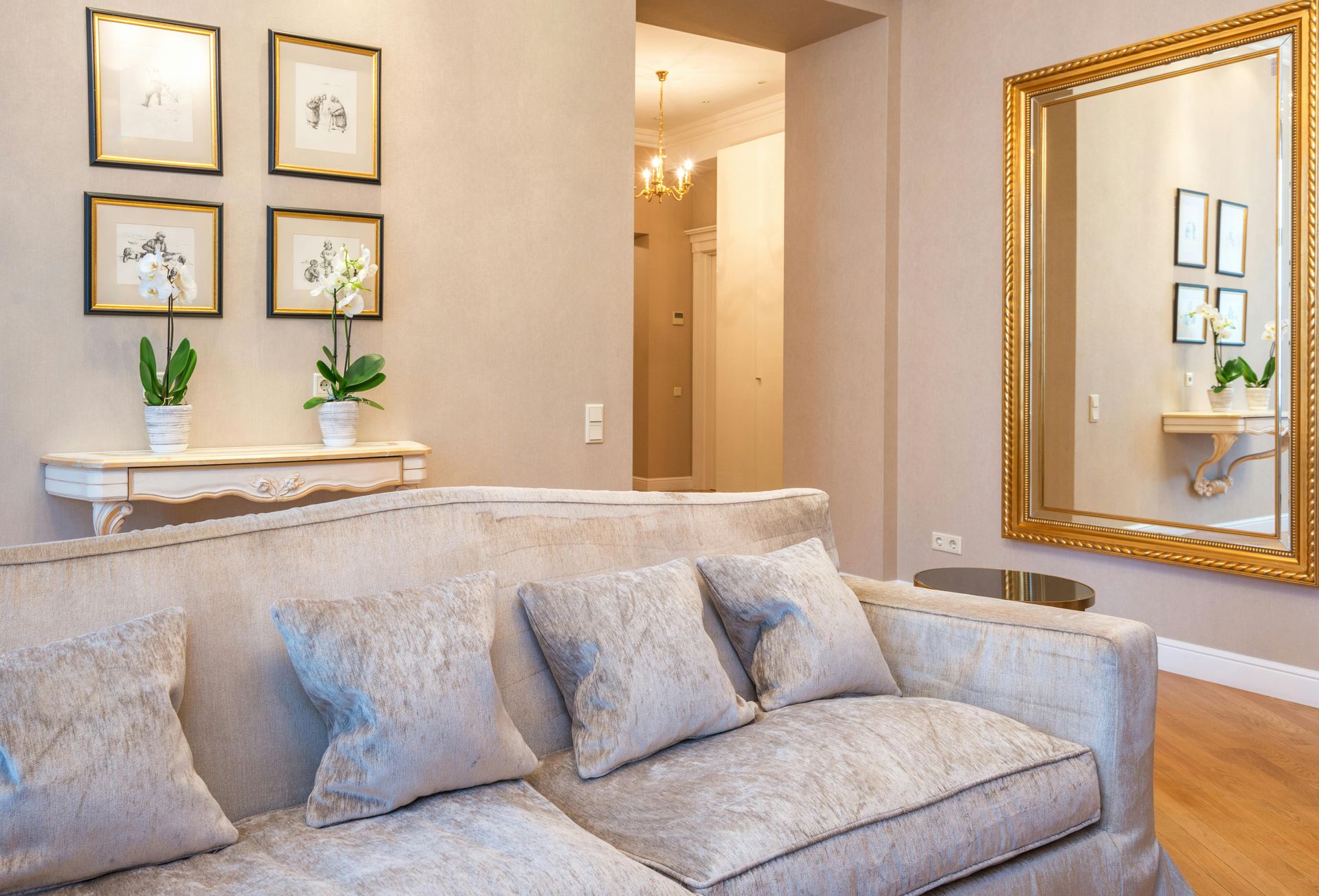 A beige sofa sits in a neutral-toned room with four framed pictures on the wall and a large gold-framed mirror nearby.