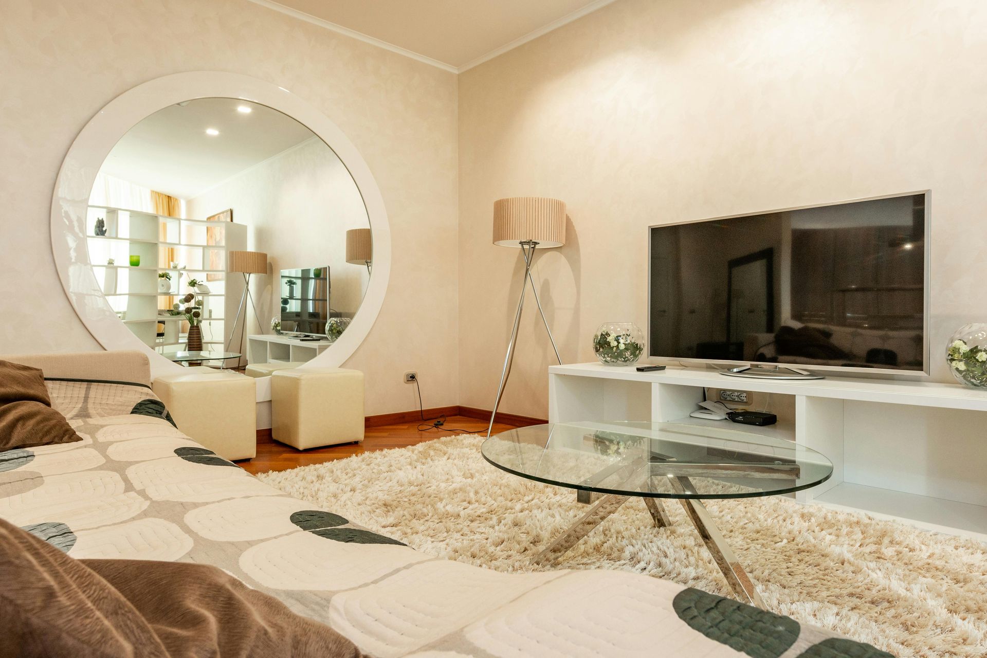 A contemporary living room with a large round mirror, a white media console, flat-screen TV, and a glass coffee table.