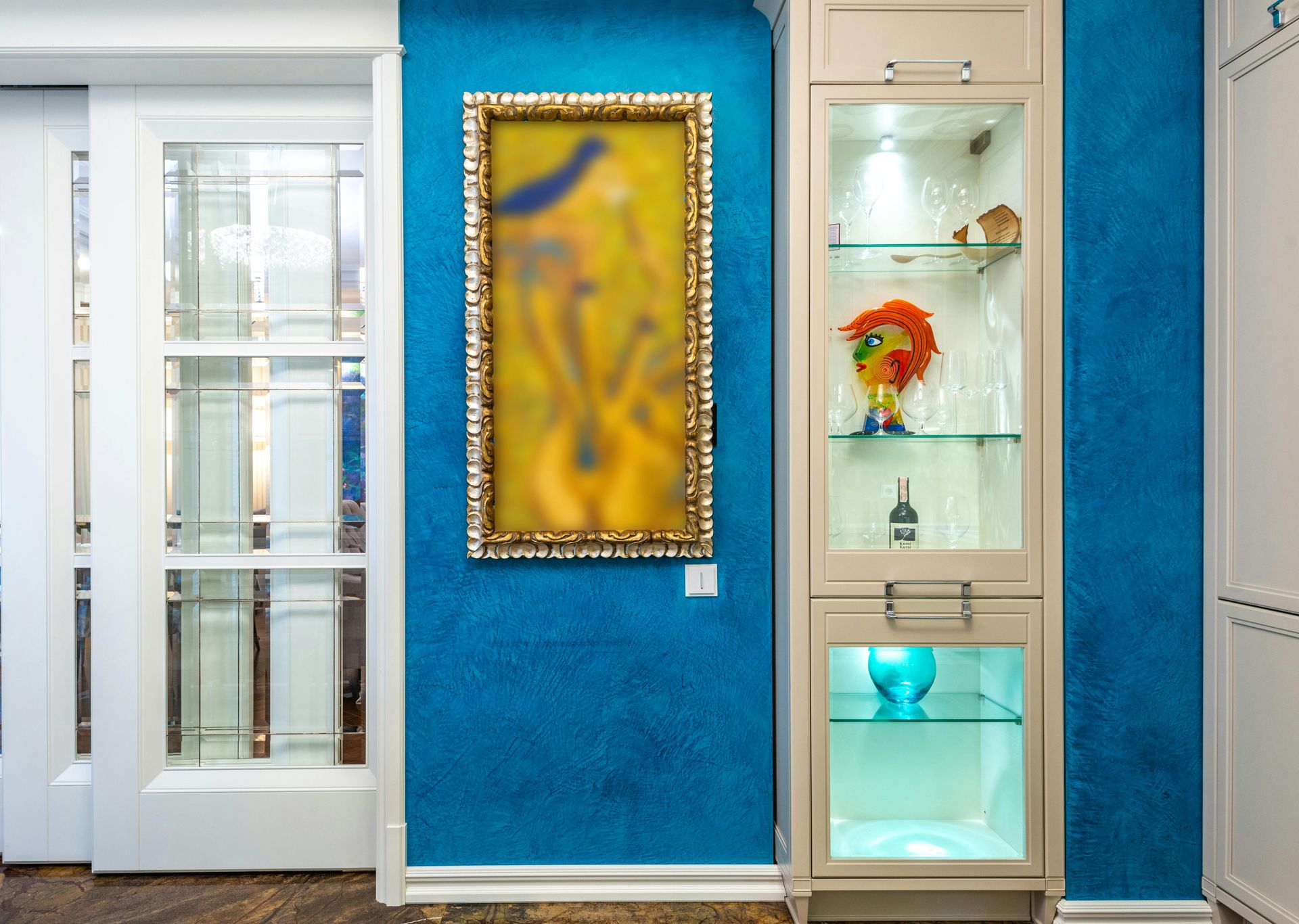 A blue wall with a framed abstract painting next to a tall, lighted, beige glass-front display cabinet.