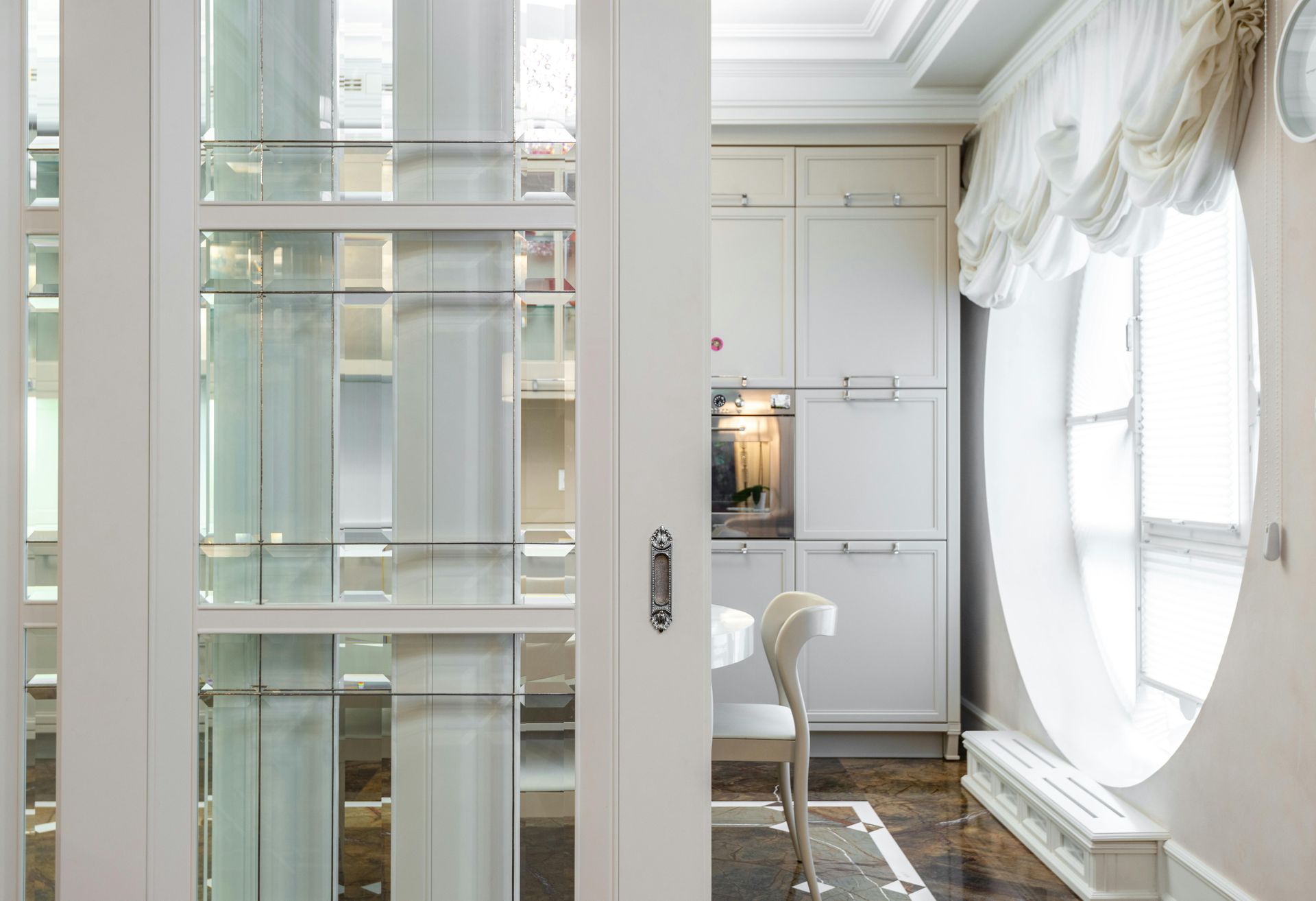 A bright room seen through a white sliding door, featuring kitchen cabinetry, a white chair, and a large circular window.