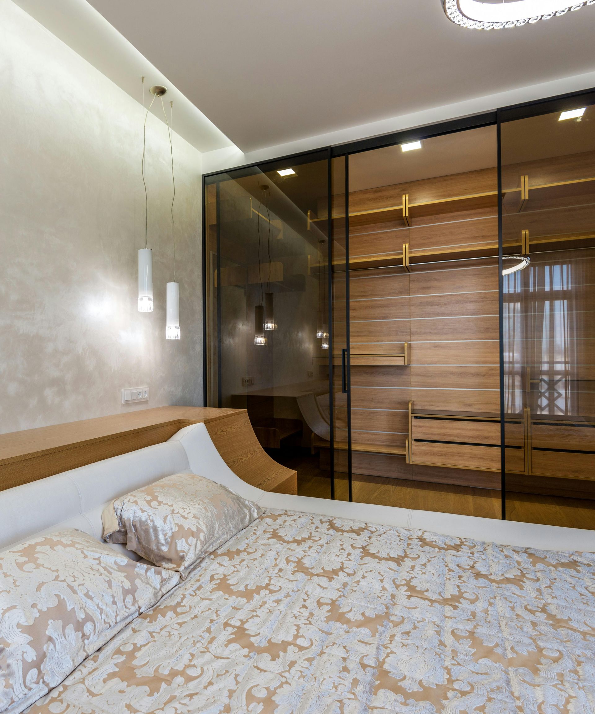 A bedroom featuring a bed with patterned bedding next to a modern walk-in closet with sliding glass doors and wood shelves.