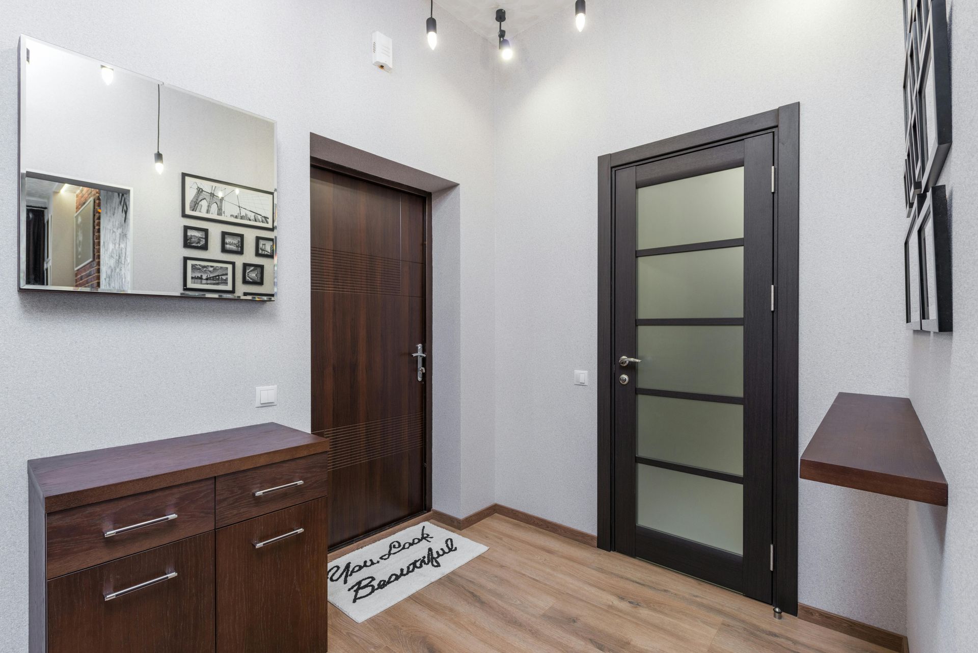 A bright hallway featuring a dark wood cabinet, a wall mirror, a dark door, a glass-paneled door, and a floating shelf.