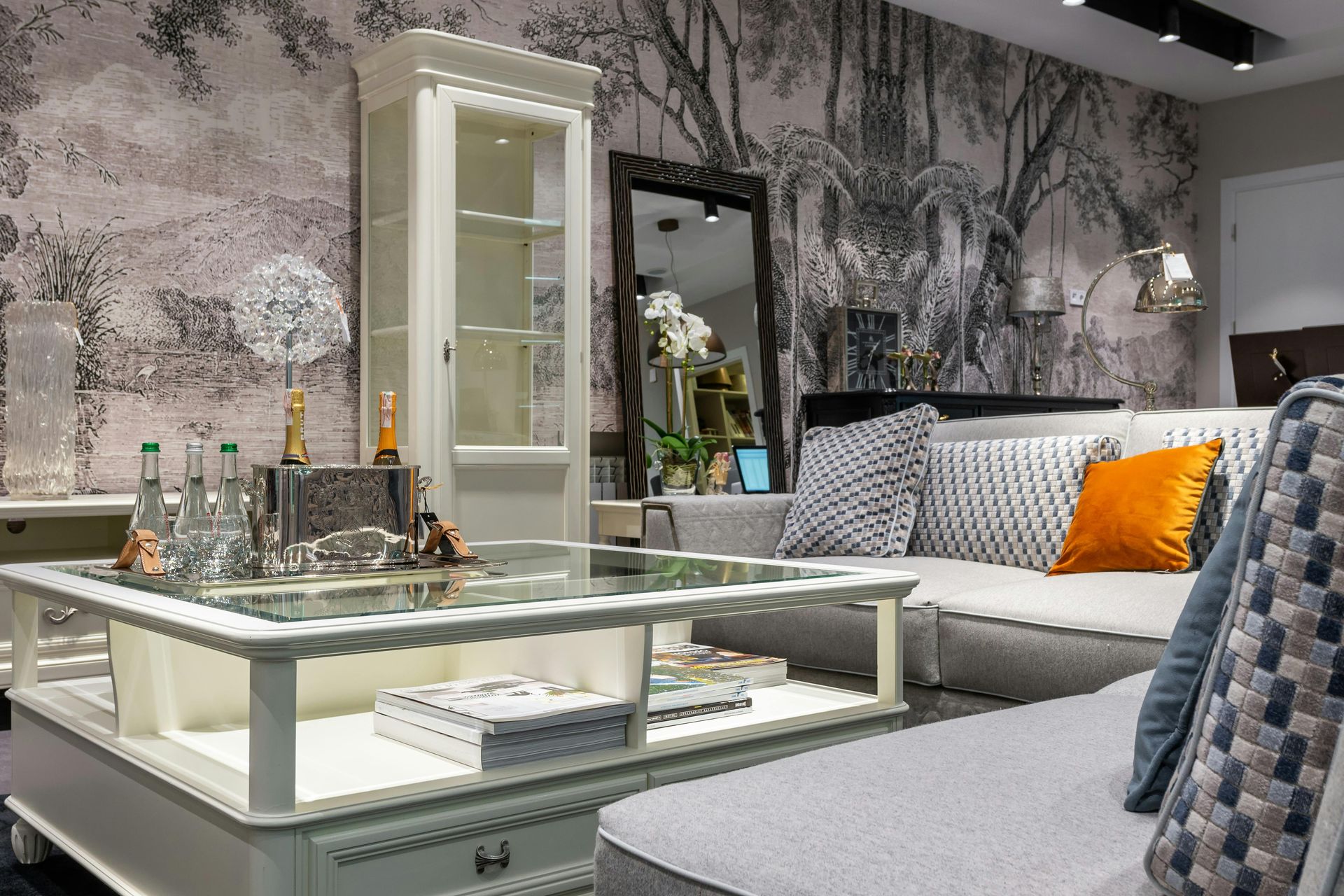 A modern living room features a glass coffee table, a grey sofa with colorful cushions, and ornate forest-print wallpaper.